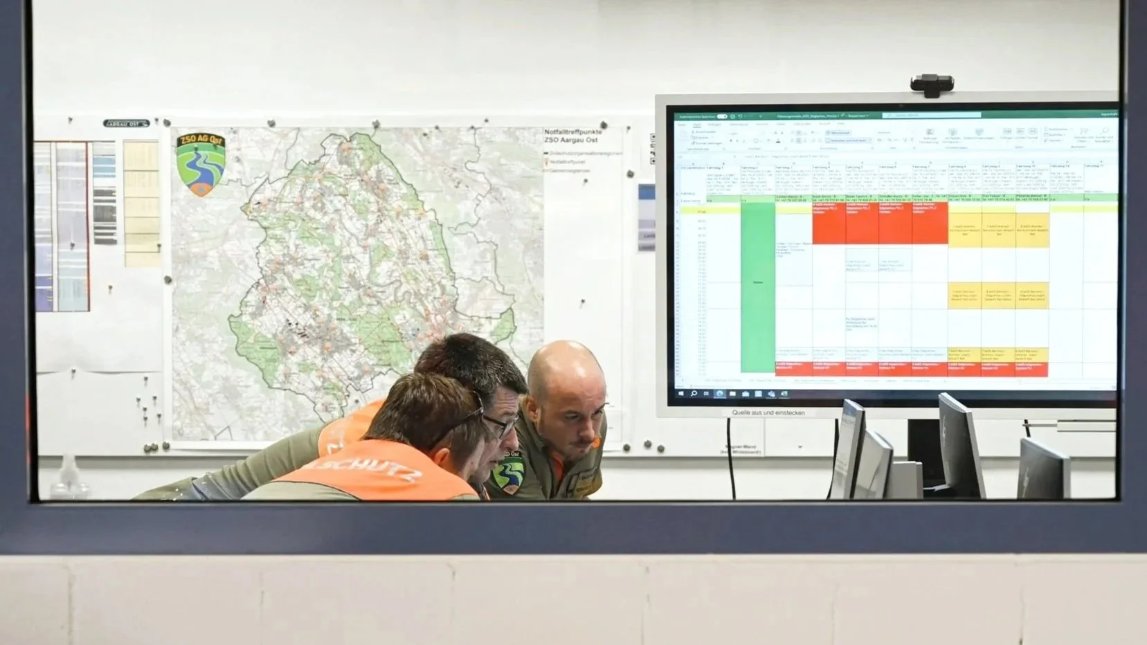 Drei Personen in einem Kontrollraum, die auf einen Bildschirm schauen, auf dem eine Tabelle mit farblich markierten Zellen und ein Stadtplan zu sehen sind.