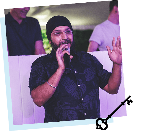 A man wearing a black turban and glasses speaking into a microphone at an event with people in the background.