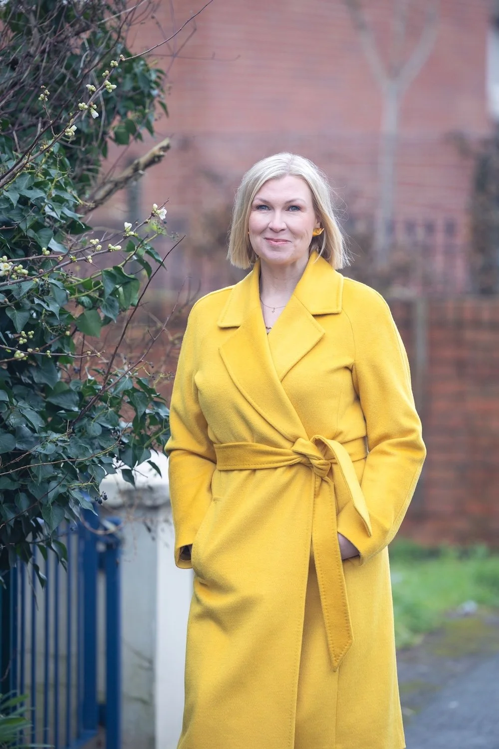 A woman with blonde hair wearing a bright yellow coat, standing outdoors near some bushes, with a brick building in the background.