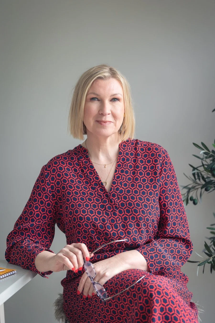 A woman with blonde hair wearing a red and navy geometric patterned dress sitting indoors with a neutral background and a plant to her right, holding sunglasses in her left hand.