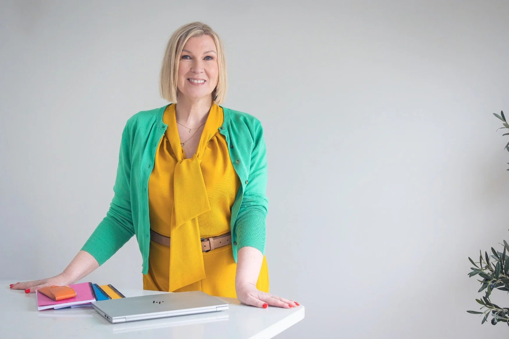 A woman with blonde hair, wearing a yellow blouse and green cardigan, leaning on a white table with colorful notebooks, a laptop, and a pink wallet, in a bright room with minimal decor.