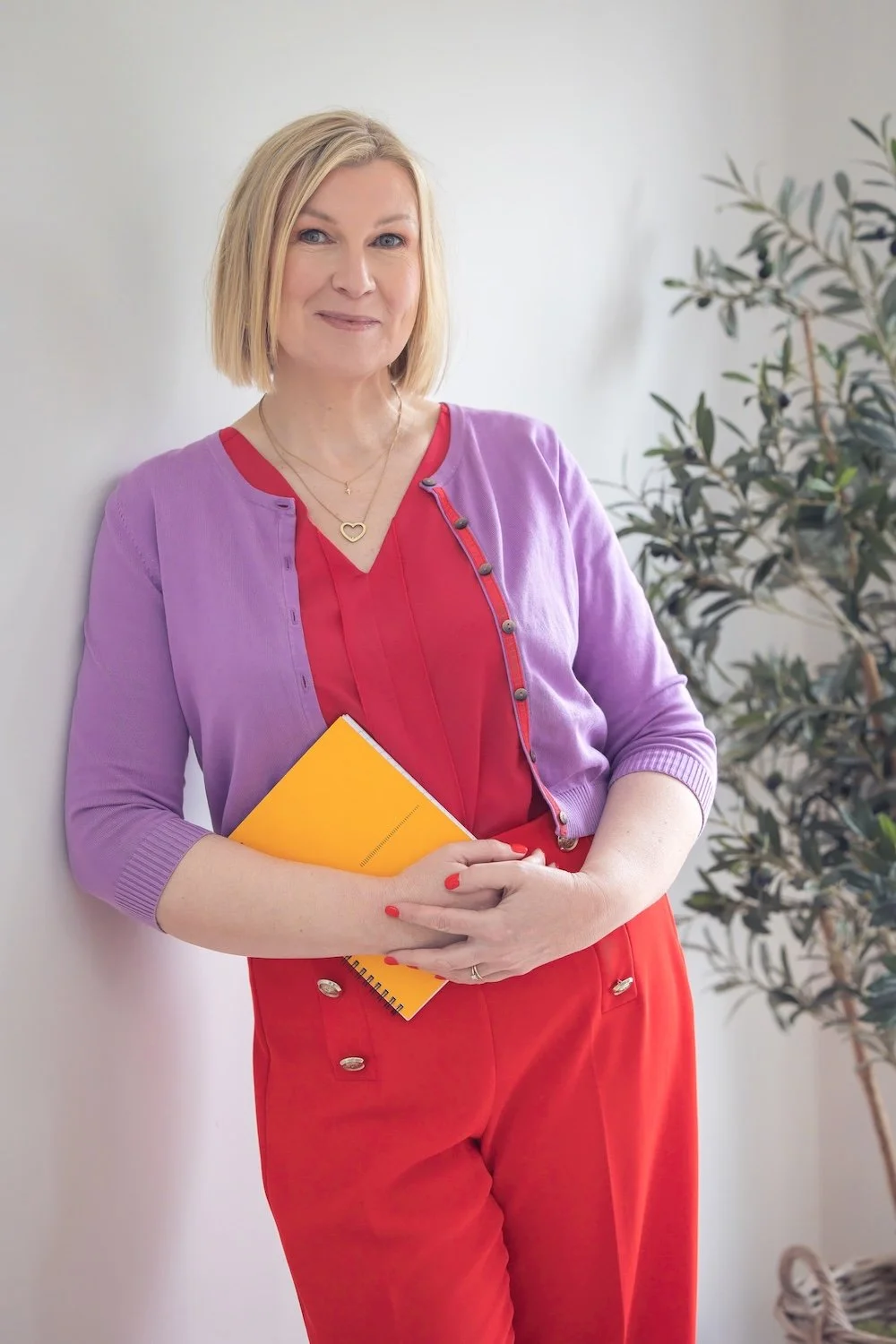 A woman with blonde hair wearing a red blouse, purple cardigan, and red pants, holding a yellow notebook, standing indoors next to a white wall and green plant.