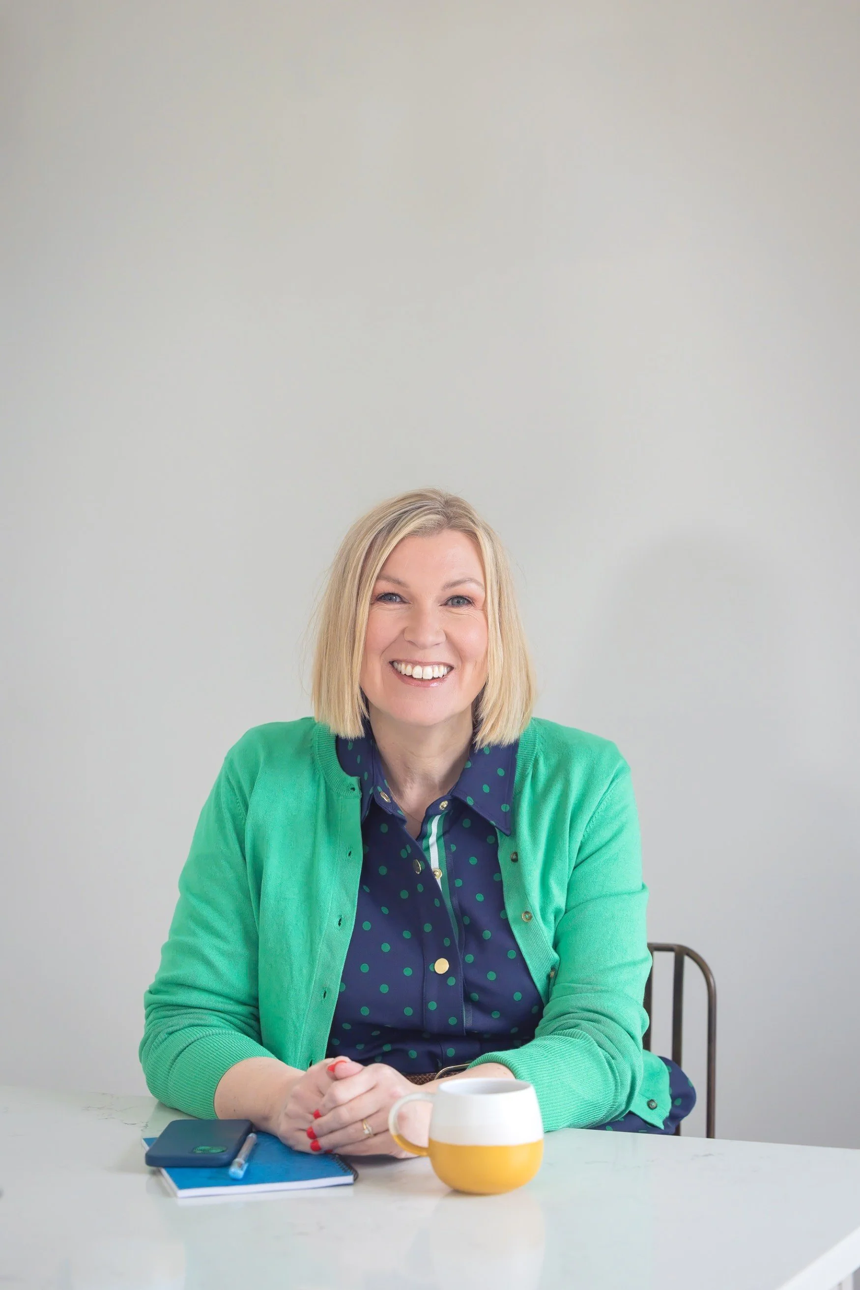 A woman with blonde hair smiling, sitting at a white table with a yellow and white mug, a closed notebook, and a smartphone. She is wearing a blue shirt with green polka dots and a green cardigan.