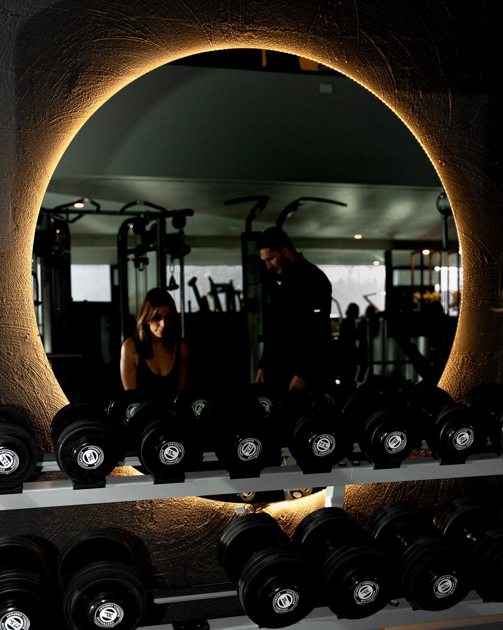 A gym scene viewed through a circular mirror showing a woman and a man with various exercise equipment visible in the background.
