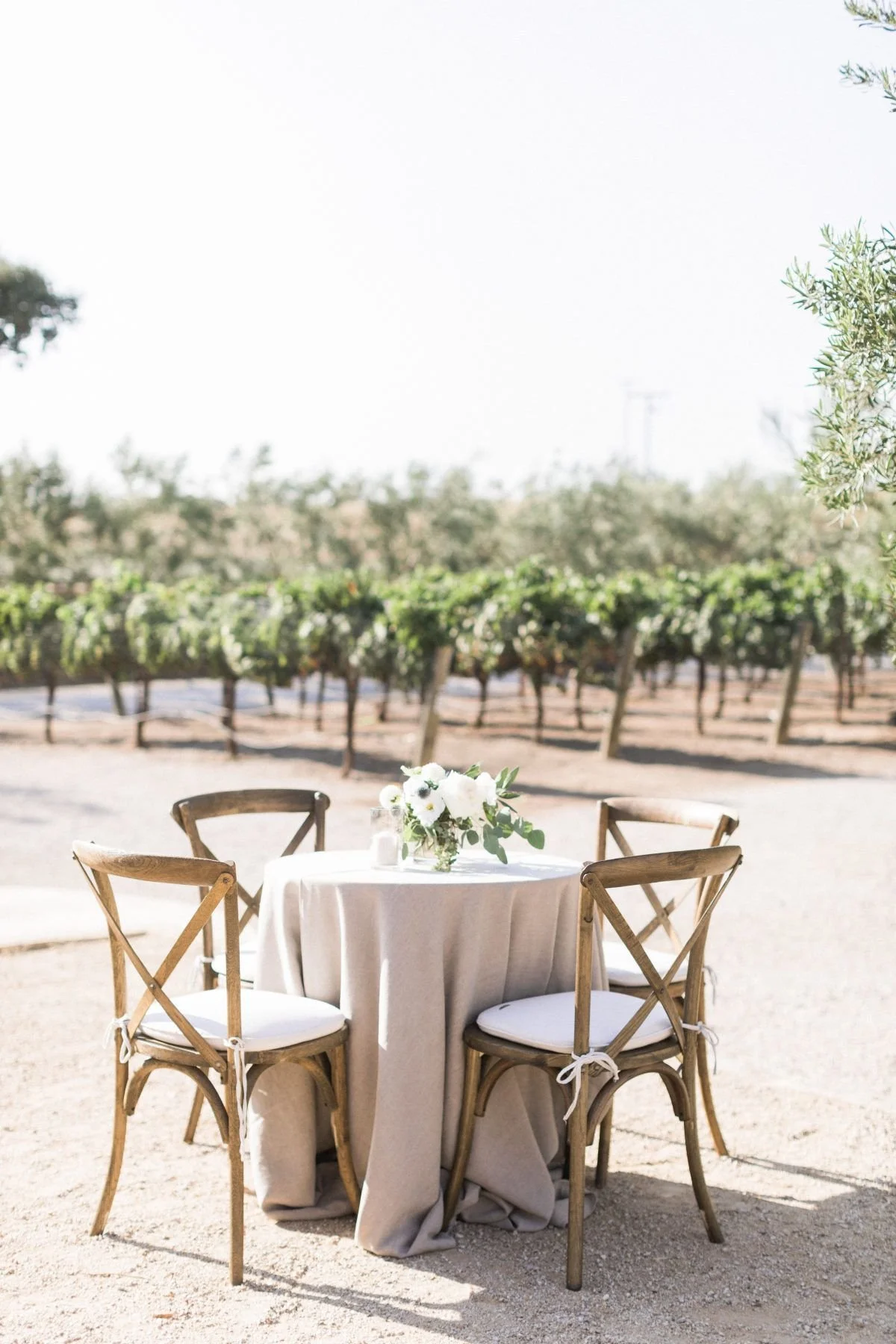 Round table covered with a white tablecloth and a floral centerpiece, surrounded by four wooden chairs with white cushions, in an outdoor vineyard setting.