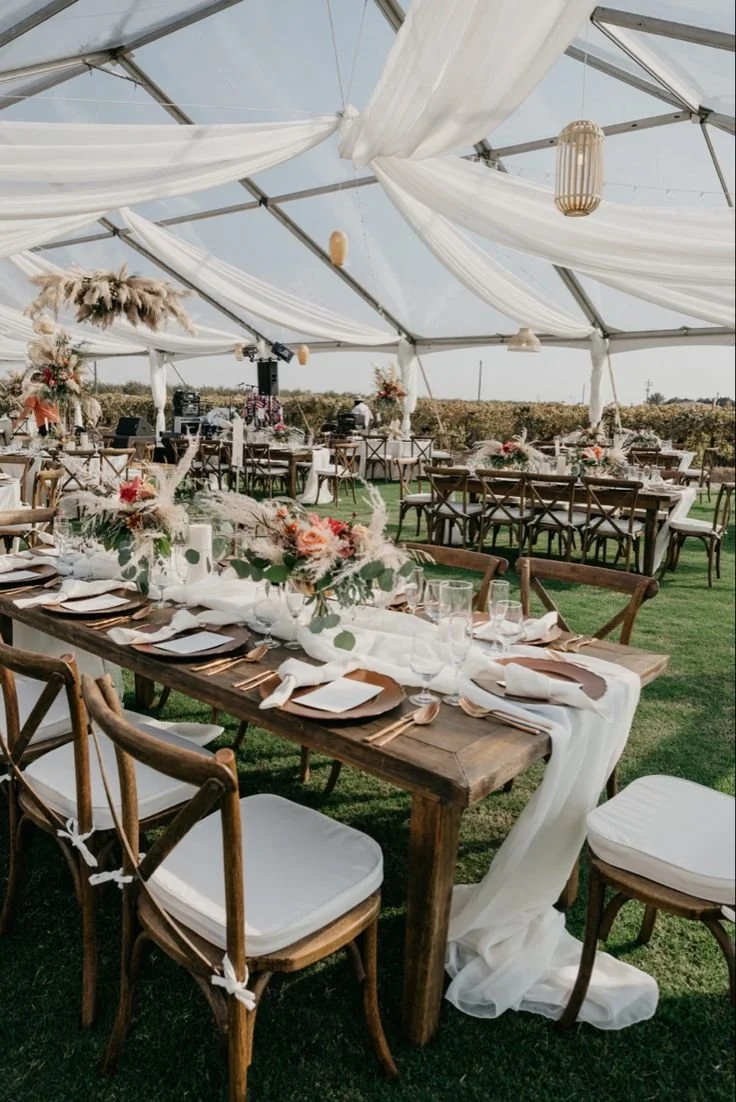 Decorated outdoor wedding reception under a tent with wooden tables, white drapery, floral centerpieces, and wooden chairs.