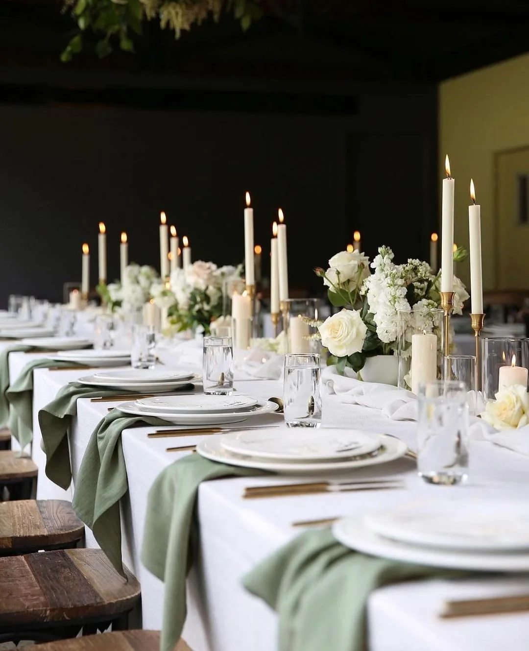 Elegant dining table set for a formal event with white tablecloths, green napkins, white plates, glassware, and floral centerpieces with white roses and candles.