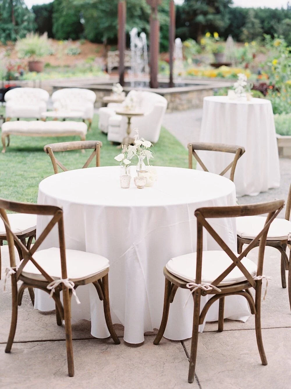 Outdoor wedding reception area with round table with white cloth, wooden chairs with white cushions, vases with white flowers, and background with seating and greenery.
