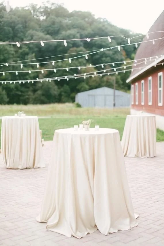 Outdoor event setup with three round tables covered in white tablecloths on a brick surface, string lights overhead, a red barn to the right, a metal shed in the background, and green trees and grass beyond.