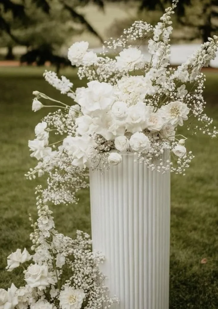 A white floral arrangement with roses and baby's breath on a white striped pedestal outdoors