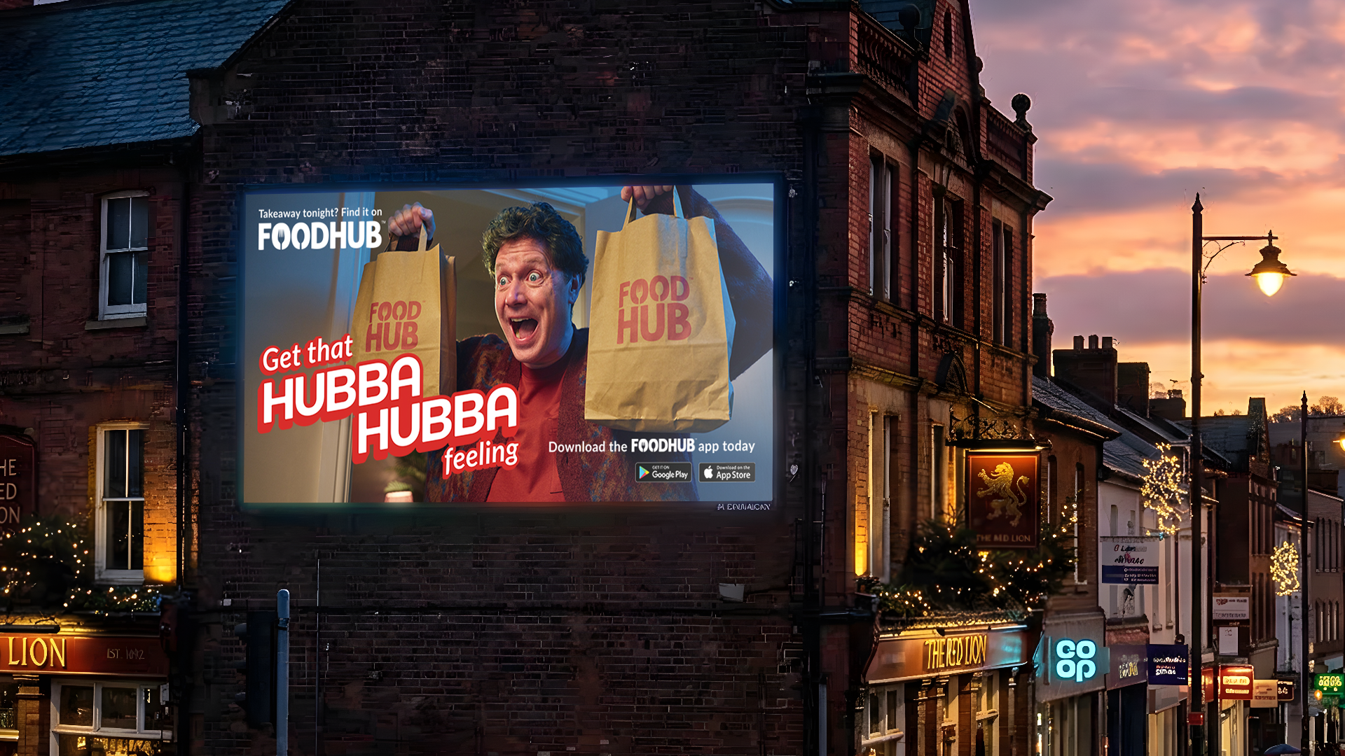 A city street during sunset with a large digital billboard advertising FOODHUB, showing a person excitedly holding shopping bags with FOODHUB branding, and text promoting the FOODHUB app. The street has lighting fixtures, shops, and holiday decorations.