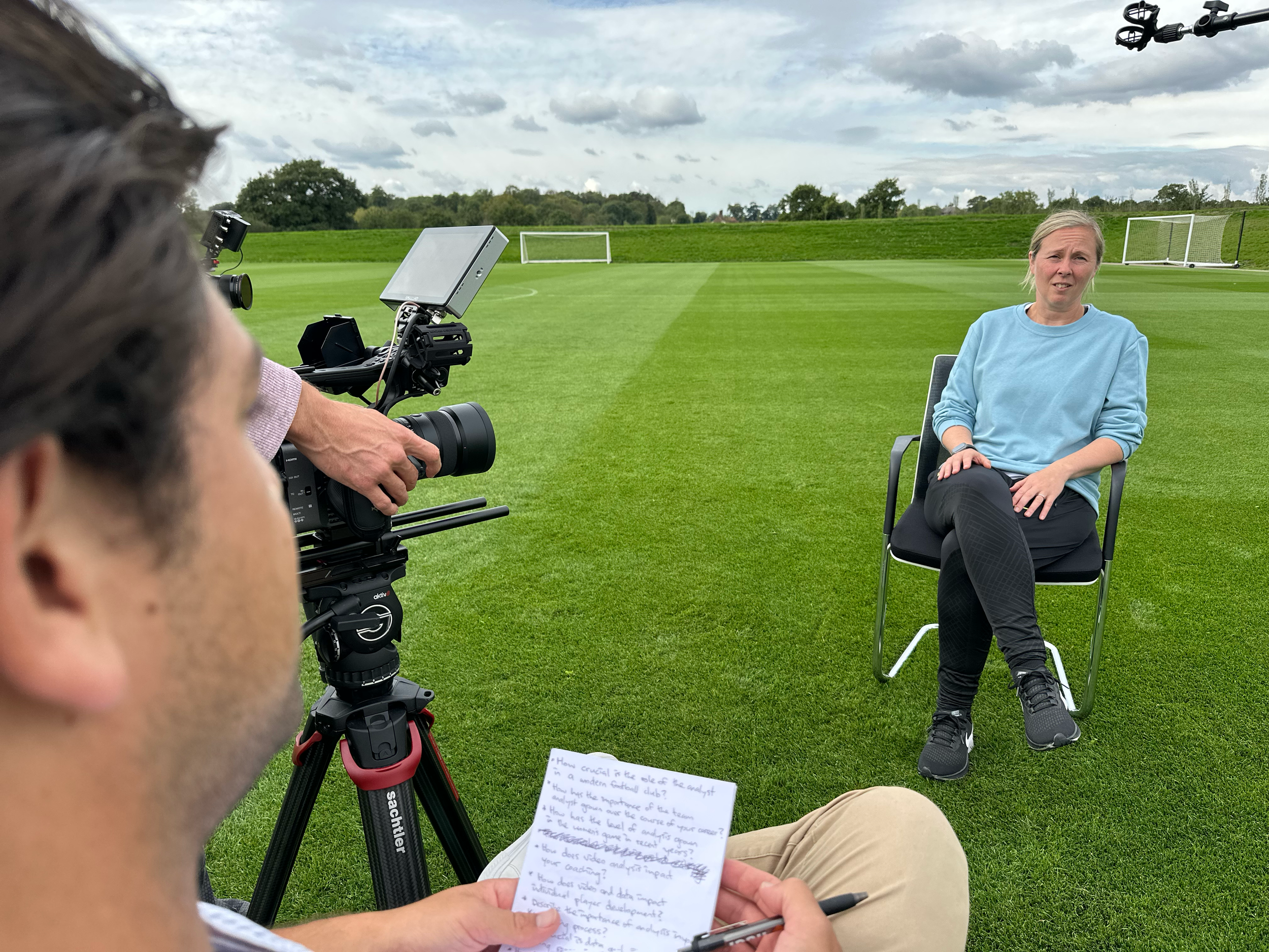 A woman sitting on a chair outdoors on a grassy field during the day being filmed by a camera crew. The woman appears to be speaking or being interviewed, and the crew is recording her.