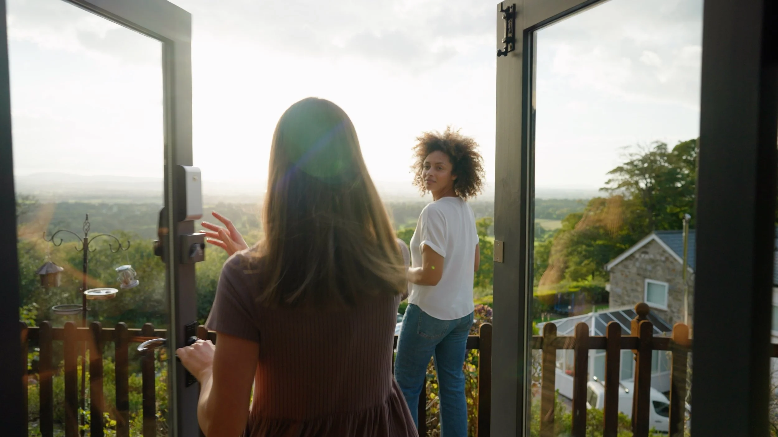 Two women are standing on a balcony, looking out at a green landscape with trees and houses in the distance. The woman closer to the camera is partially obscured, with her back to the camera, and the woman facing the camera is wearing a white t-shirt and jeans. The scene is bright, with sunlight creating a lens flare.