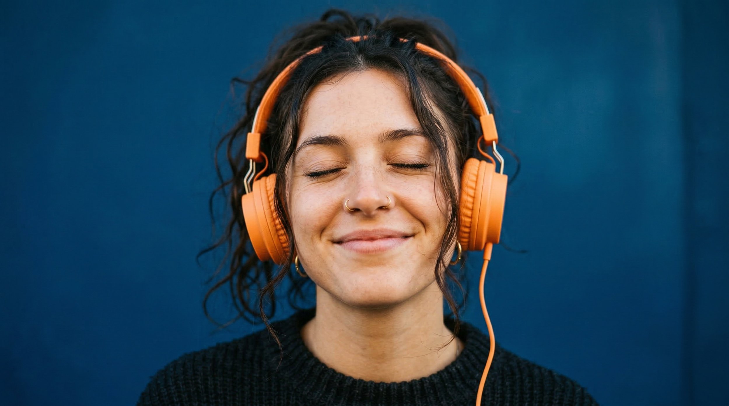 A woman with curly dark hair wearing orange headphones with closed eyes and a serene smile, standing against a dark blue background.