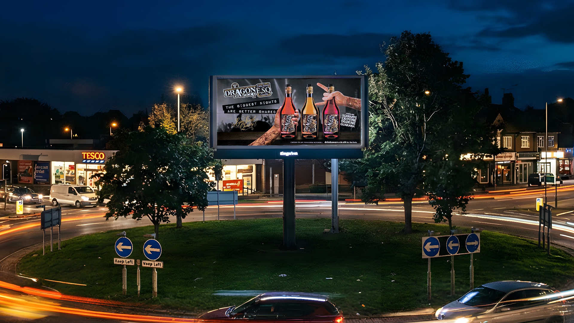 Nighttime scene of a traffic roundabout with a large billboard advertising drinks, trees, cars, and a Tesco store in the background.