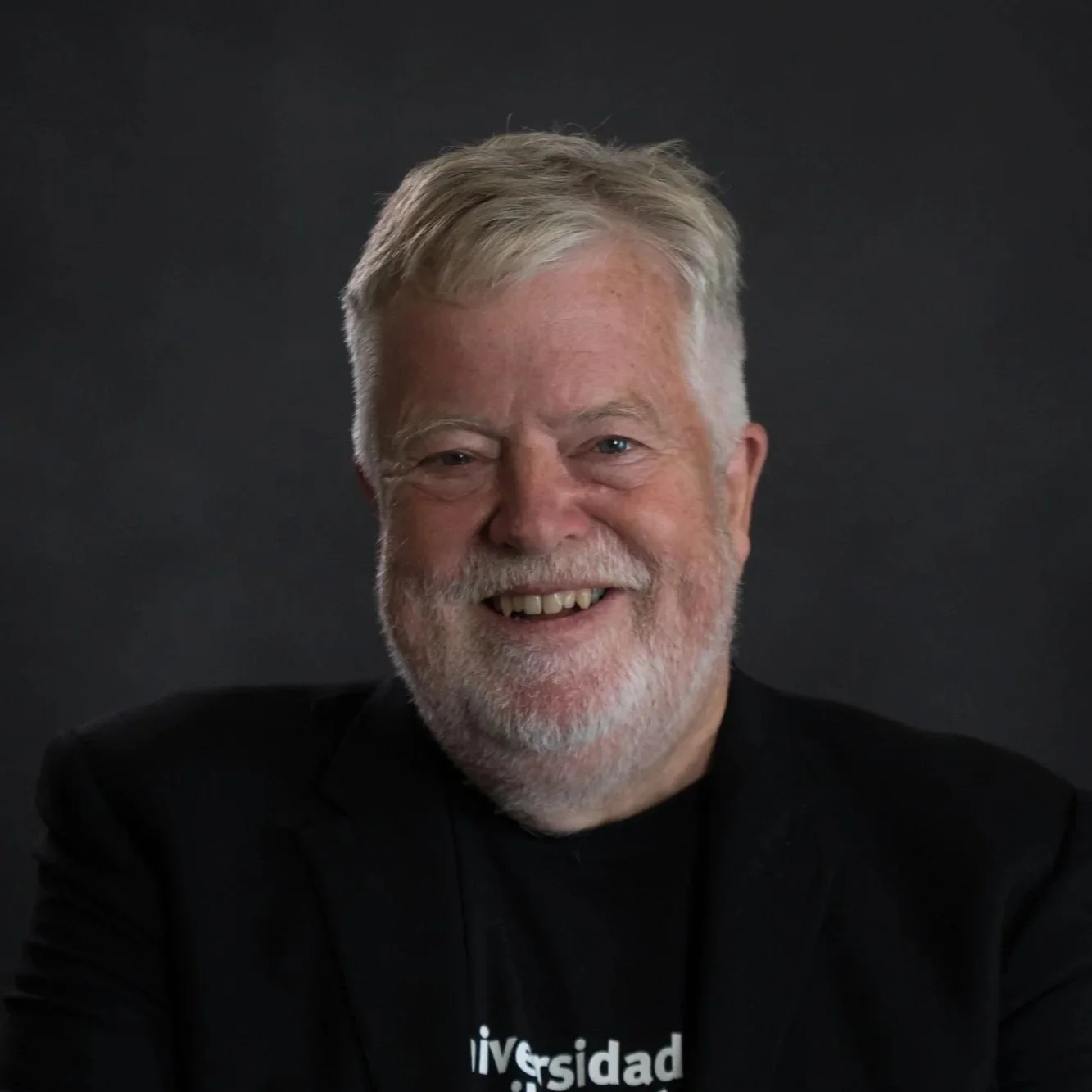 Proudly sporting a black Universidad Camilo Jose Cela t-shirt (Stephen is chair of Learning and Innovation) under a black blazer, Stephen's warm smile stands out against the dark background behind