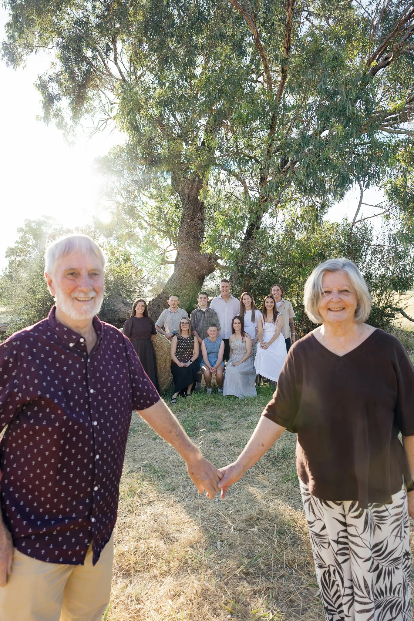 An elderly man and woman holding hands in the foreground, smiling, with a group of younger people and a large tree in the background during sunset.