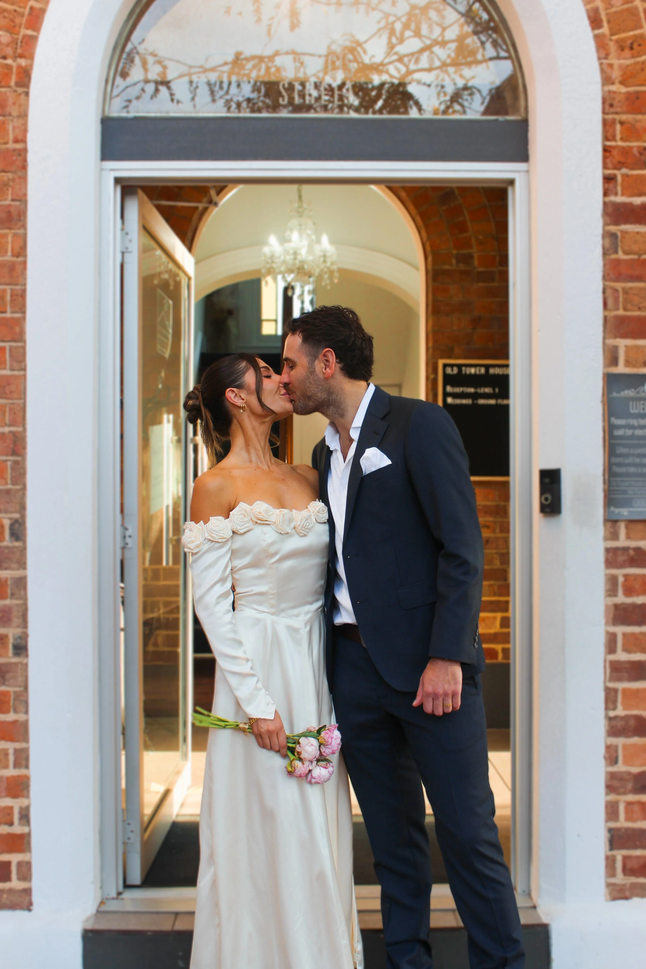 A couple dressed in wedding attire sharing a kiss at the entrance of a building with brick walls and an arched doorway.