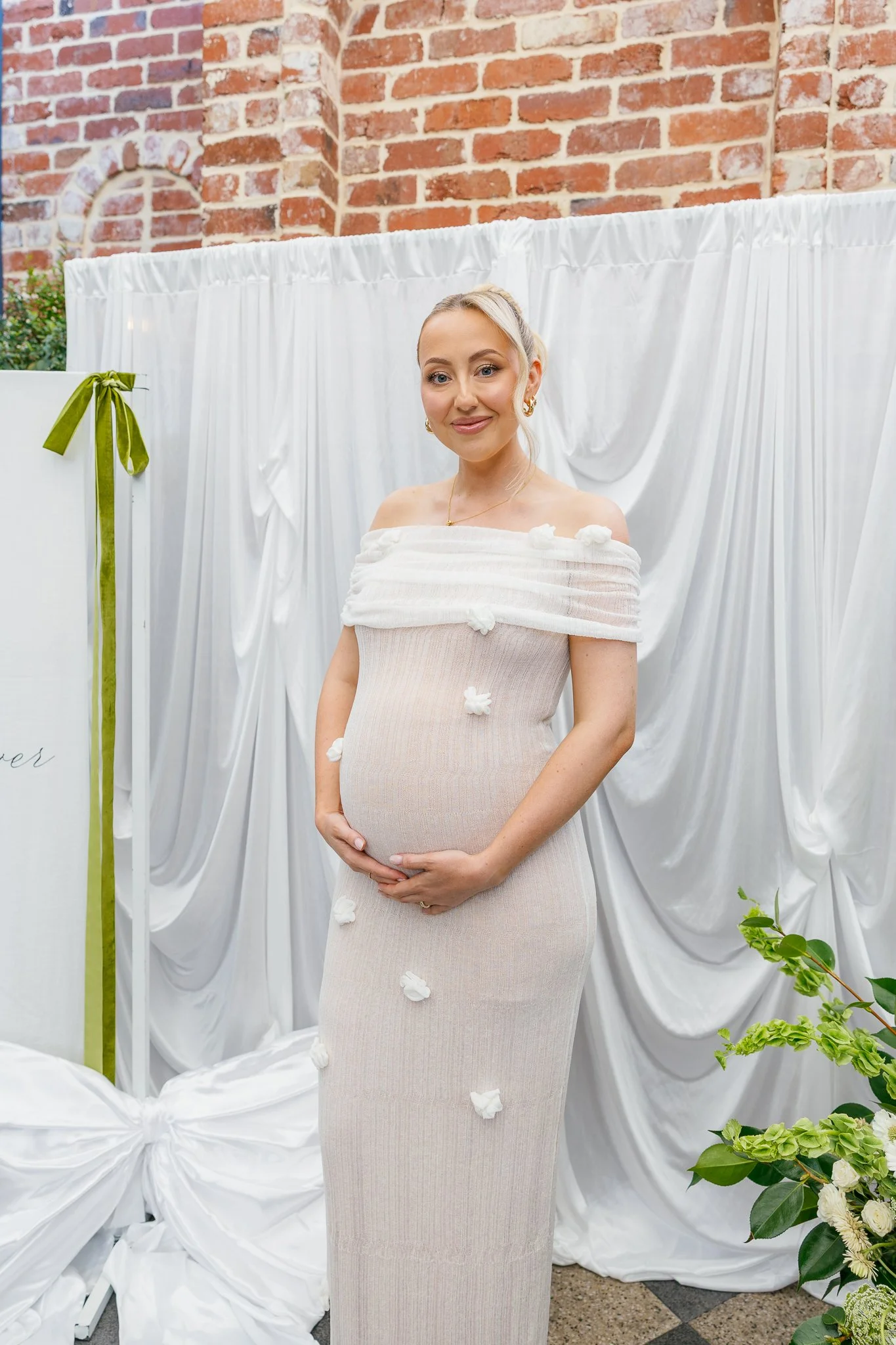 A pregnant woman in a beige dress, standing in front of white drapery and brick wall, smiling and holding her belly.