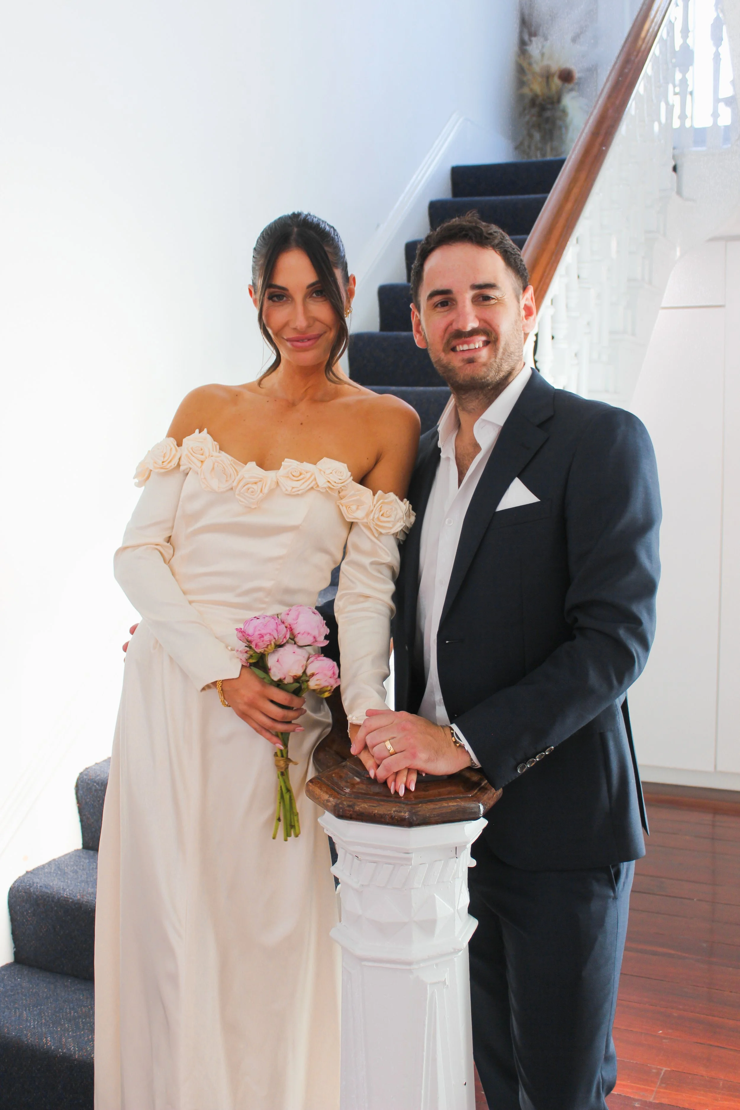 A woman in an off-shoulder wedding dress holding pink roses and a man in a dark suit with a white shirt standing together on a staircase.
