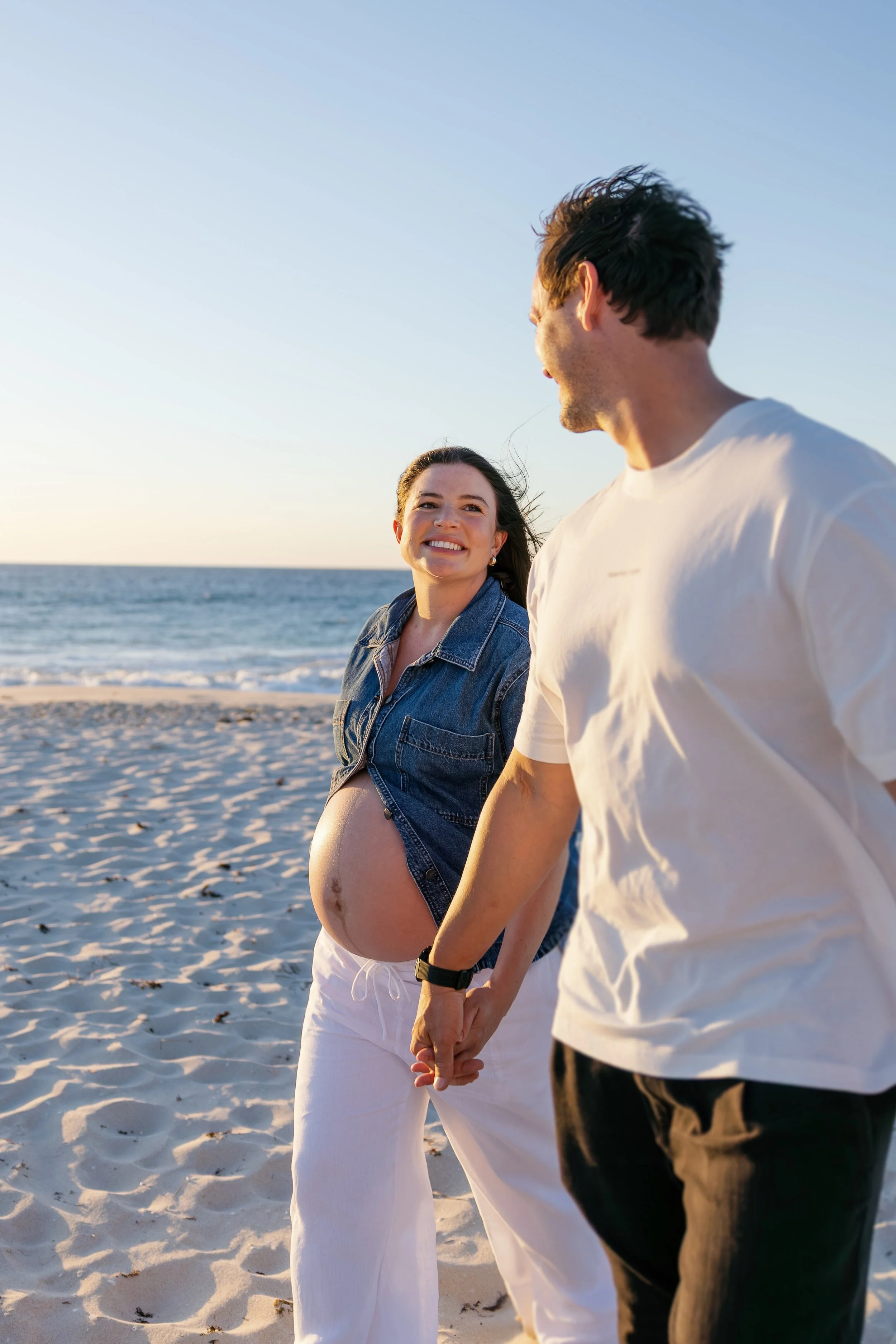 A pregnant woman and her partner hold hands and walk on the beach during sunset, with the ocean in the background.