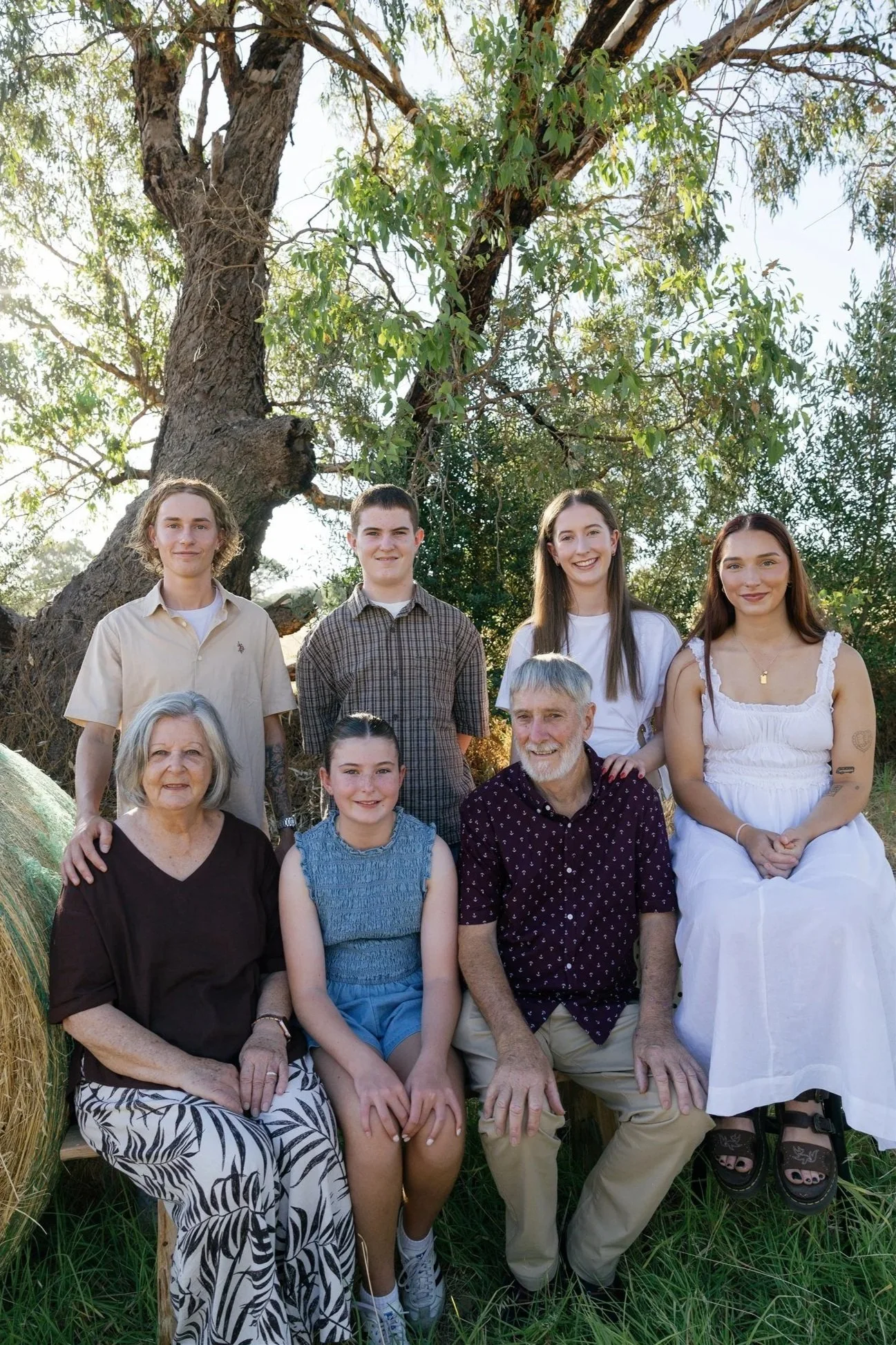 A multigenerational family portrait outdoors, featuring eight people of different ages, seated and standing in front of a large tree with green foliage, sunlight filtering through the leaves.