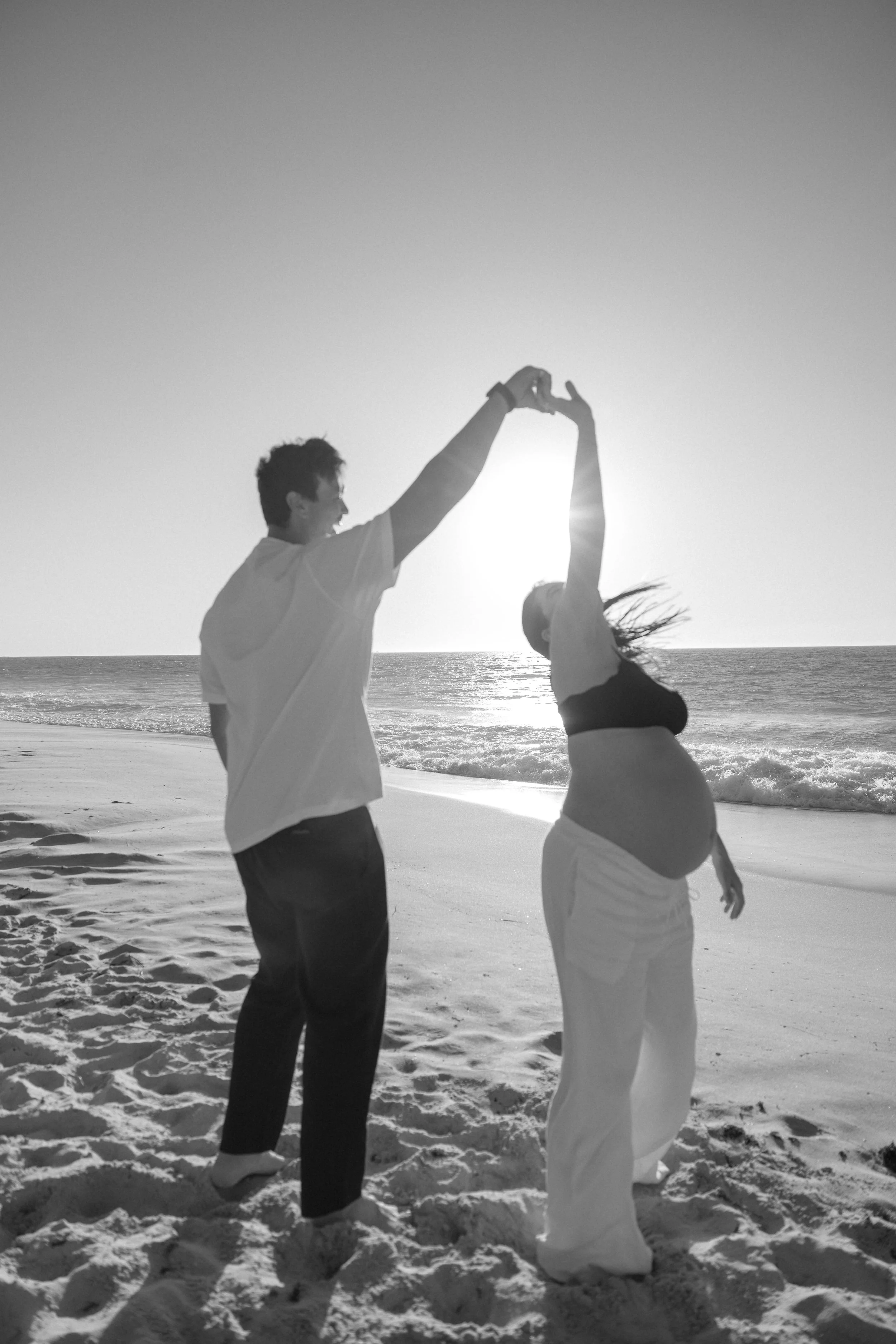 A silhouette of a pregnant woman and a man dancing on the beach at sunset, holding hands in the air.