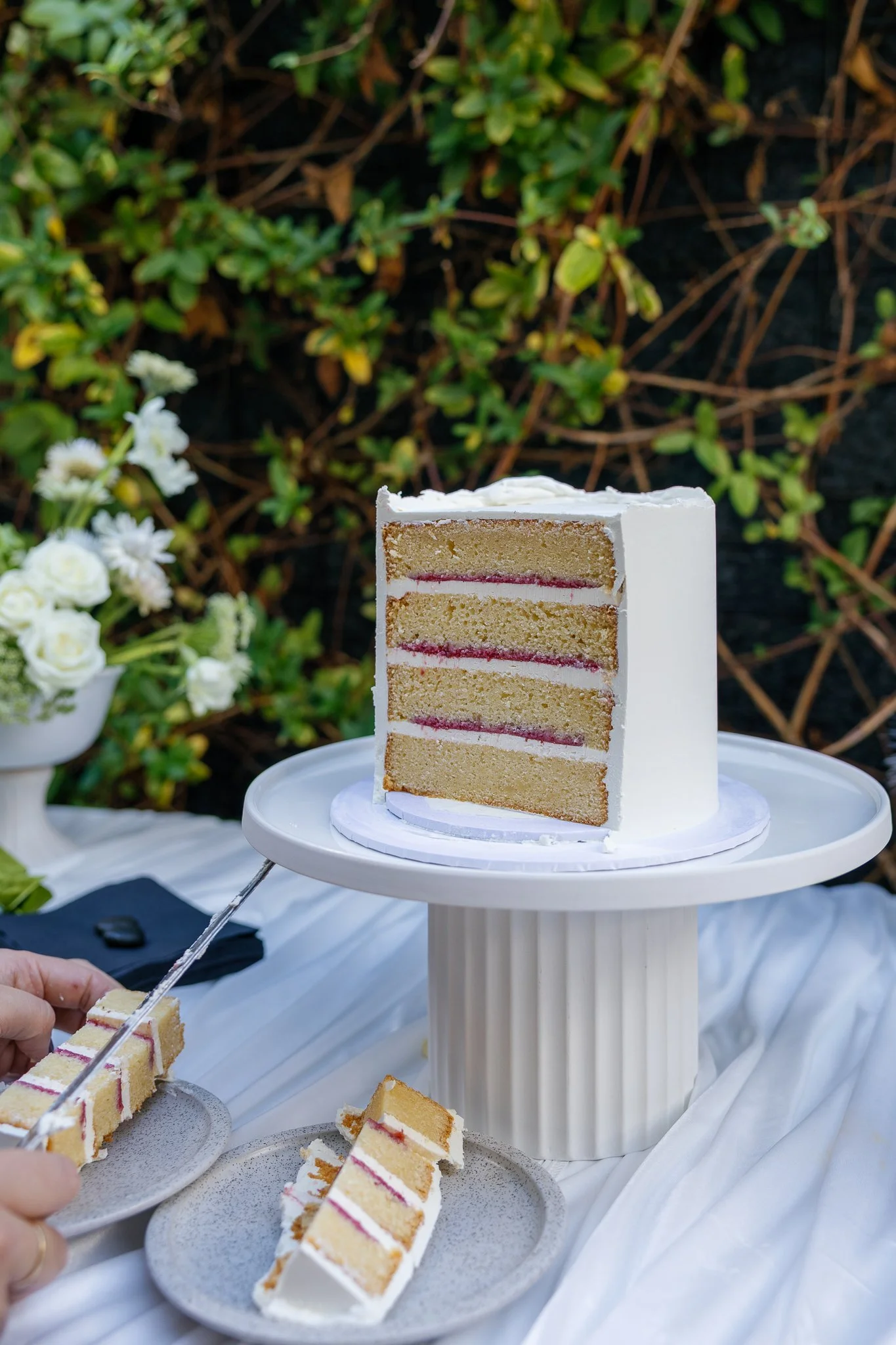 A slice of layered vanilla cake with pink filling on a white cake stand, with some slices on small gray plates, outdoors with greenery in the background.
