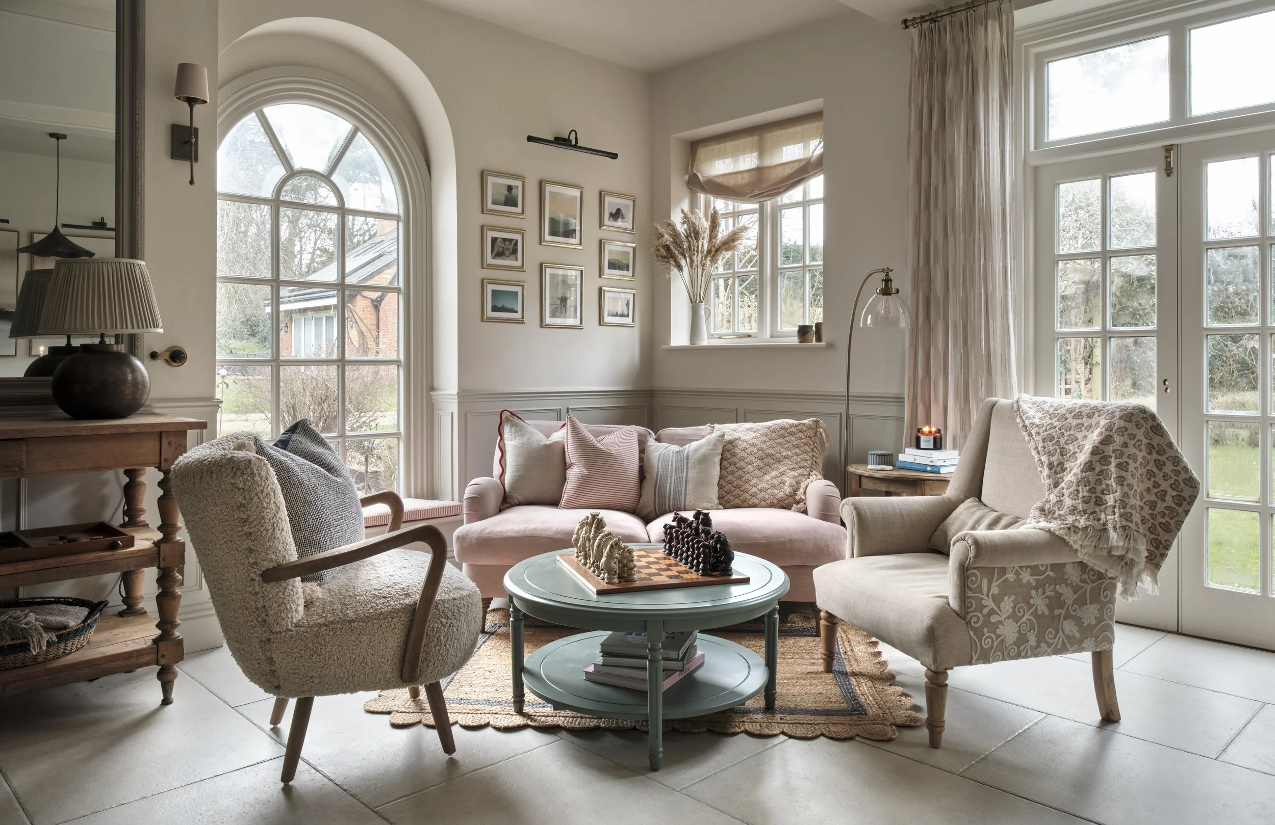 Living room with a pink sofa, beige armchair, wooden side table, and a teal coffee table with a chess set, surrounded by large windows with cream curtains and decorative pillows.