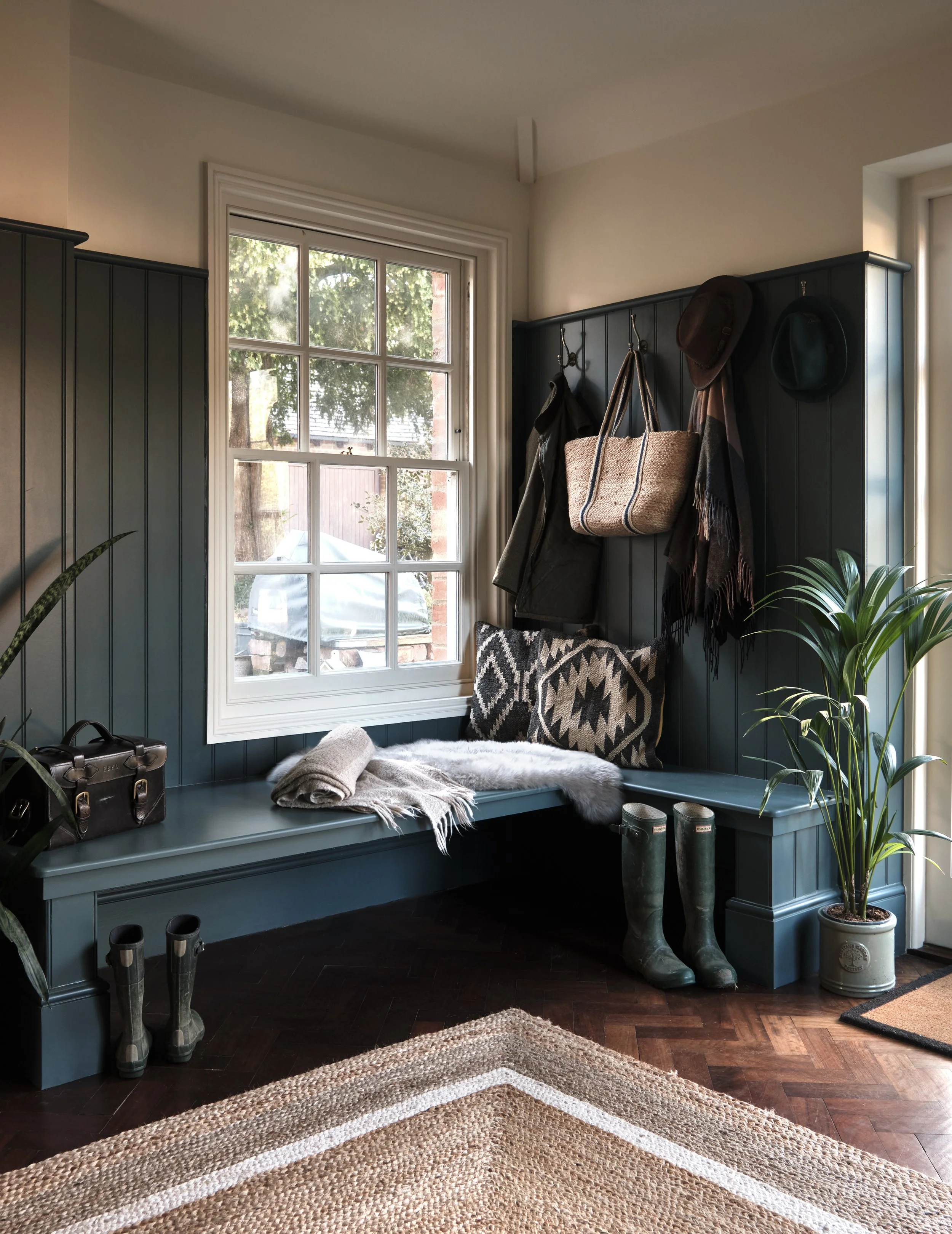 Cosy boot room with a built-in bench, coat hooks, boots, a patterned pillow, and a potted plant by a window.