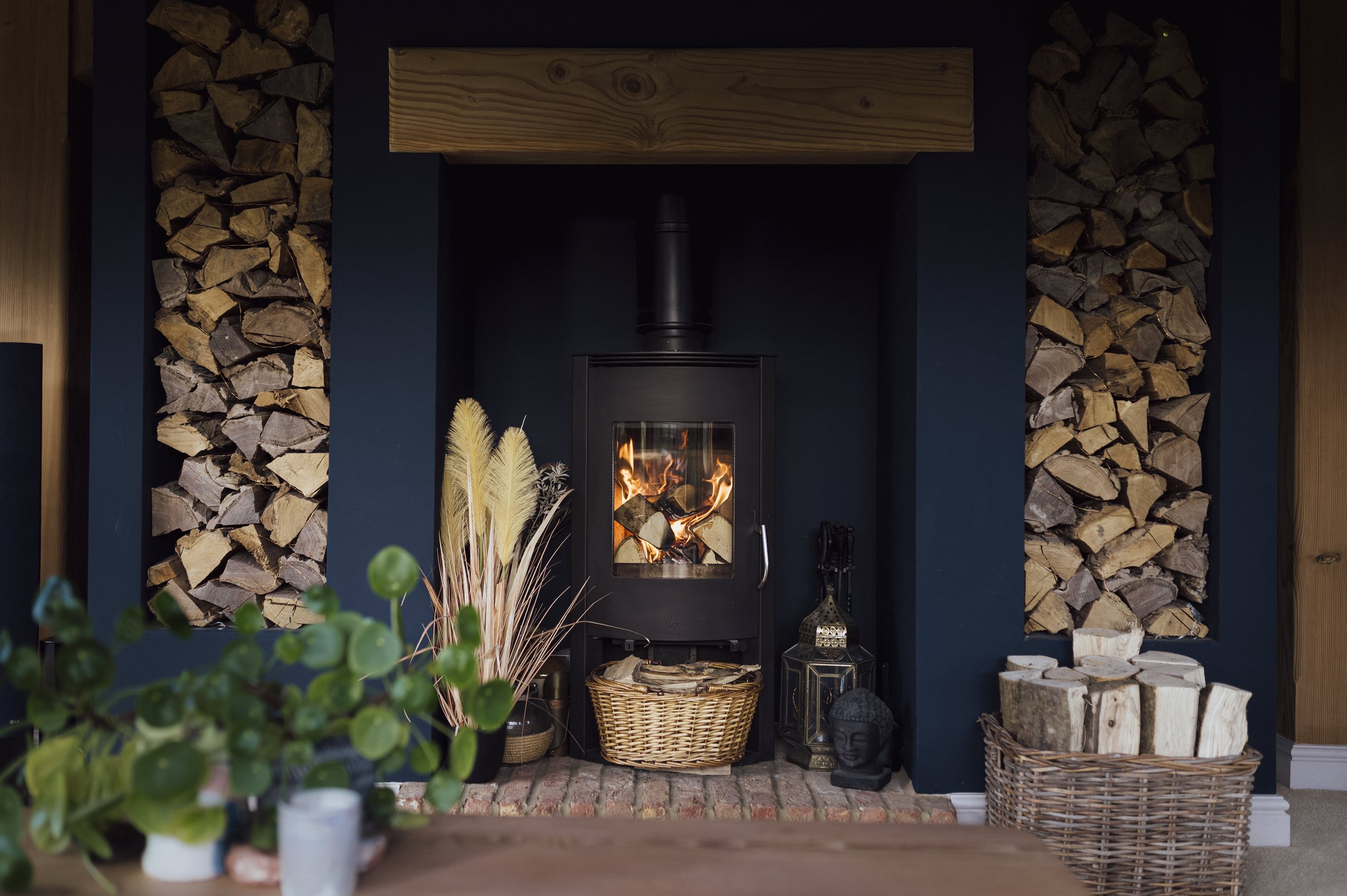 An intimate, dark interior in a deep blue colour featuring a black wood-burning stove lit with a fire, surrounded by neatly stacked firewood on both sides, decorative plants, and ambient decorative elements.