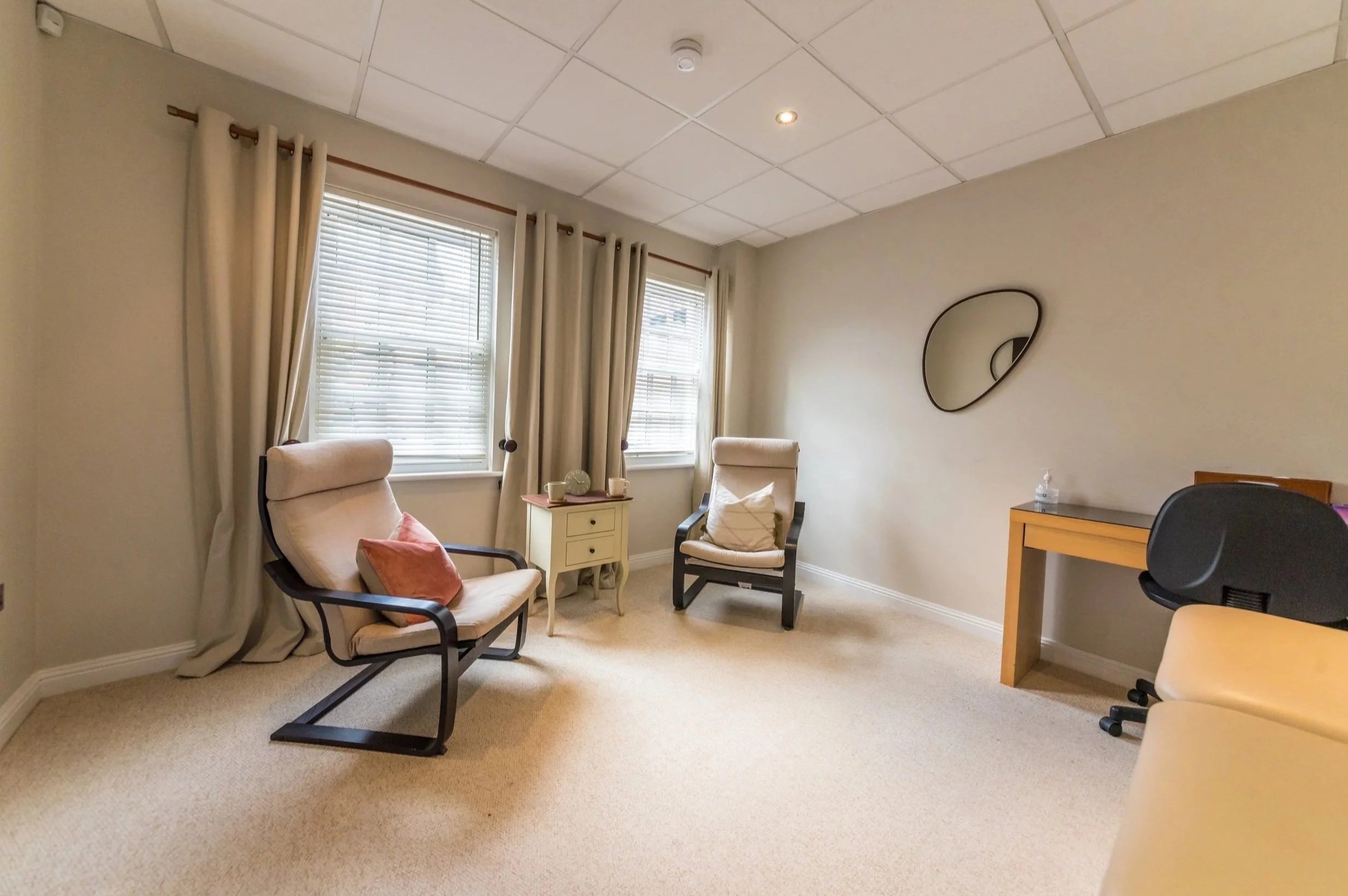 A cozy sitting area with two armchairs, one adorned with a pillow, next to a small side table with candles and a decorative item. There are three large windows with blinds and curtains, a mirror on the wall, and a desk with an office chair.