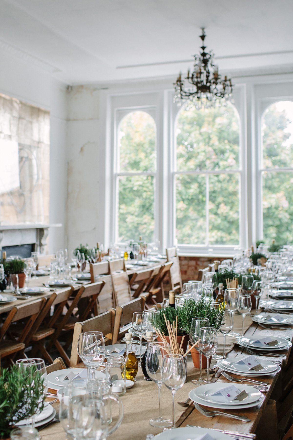 Elegant dining room setup with long wooden tables, neatly arranged with white plates, glasses, cutlery, wine bottles, and potted plants as centerpieces. Large windows provide natural light, and a chandelier hangs from the ceiling.