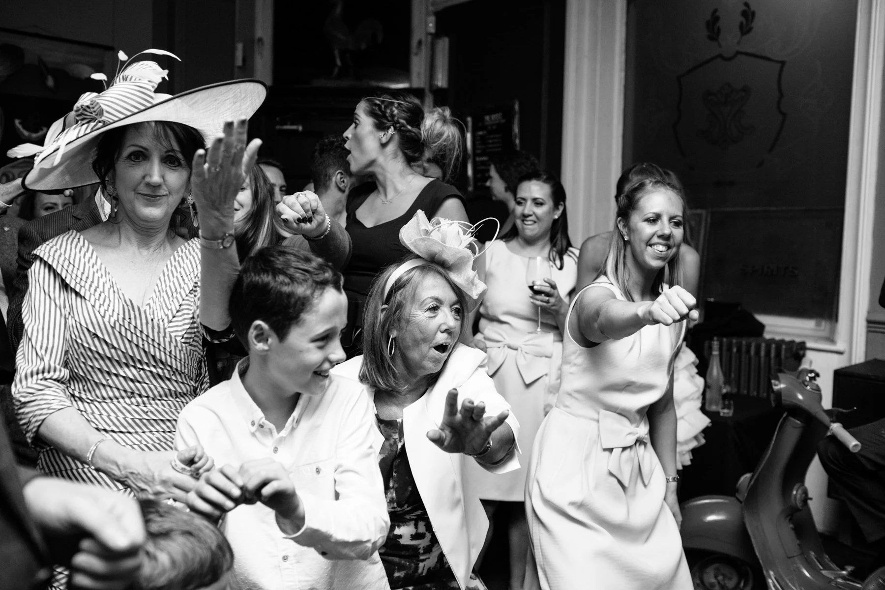 A group of people having fun and dancing, with a woman in a large hat in the foreground and others smiling and gesturing energetically, in a lively indoor setting.