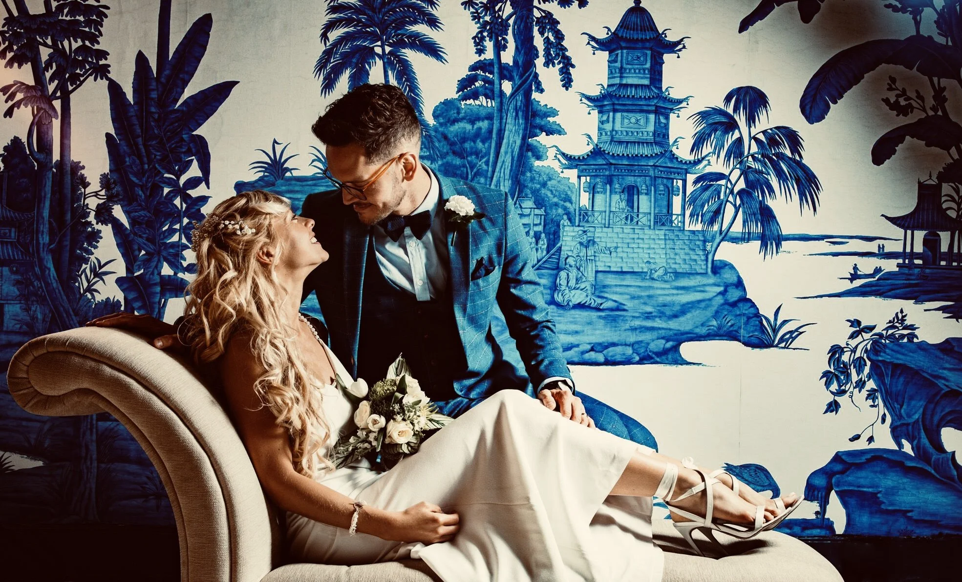 Bride and groom posing on a decorative couch with a blue and white Asian-themed mural in the background. The bride is holding a bouquet of white flowers and the groom is wearing a suit.
