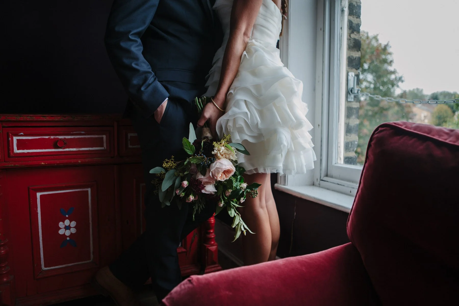 Bride and groom indoors, bride holding a bouquet, groom wearing a suit, standing near a red cabinet, window view with greenery outside.