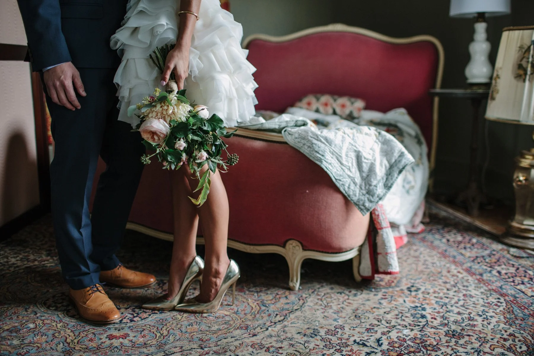 Bride in white dress holding bouquet, standing with groom in suit, near antique bed.