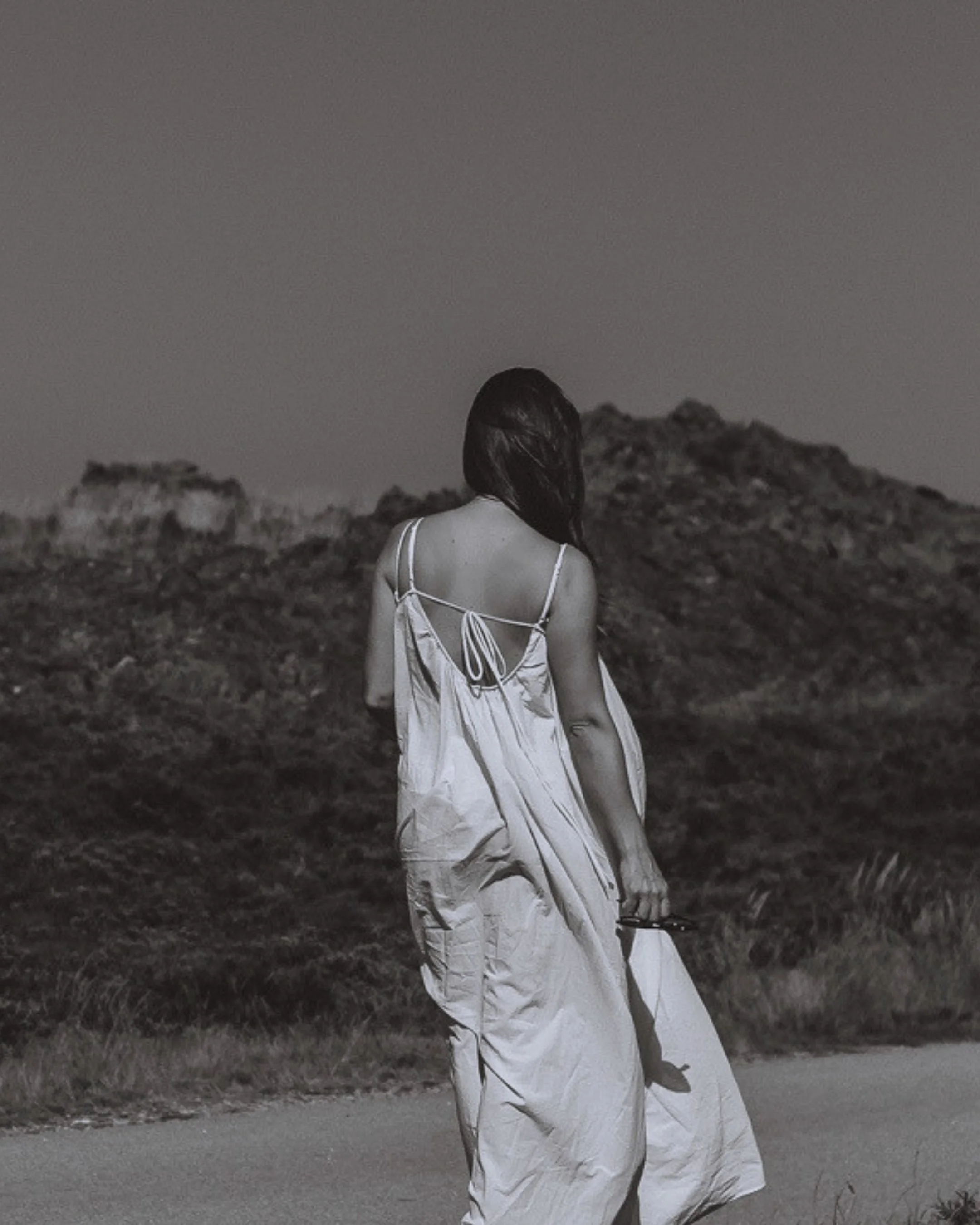 Mulher de costas usando vestido branco, com cabelo castanho escuro, caminhando em ambiente ao ar livre com terreno rochoso e vegetação ao fundo, por volta do entardecer ou noite.