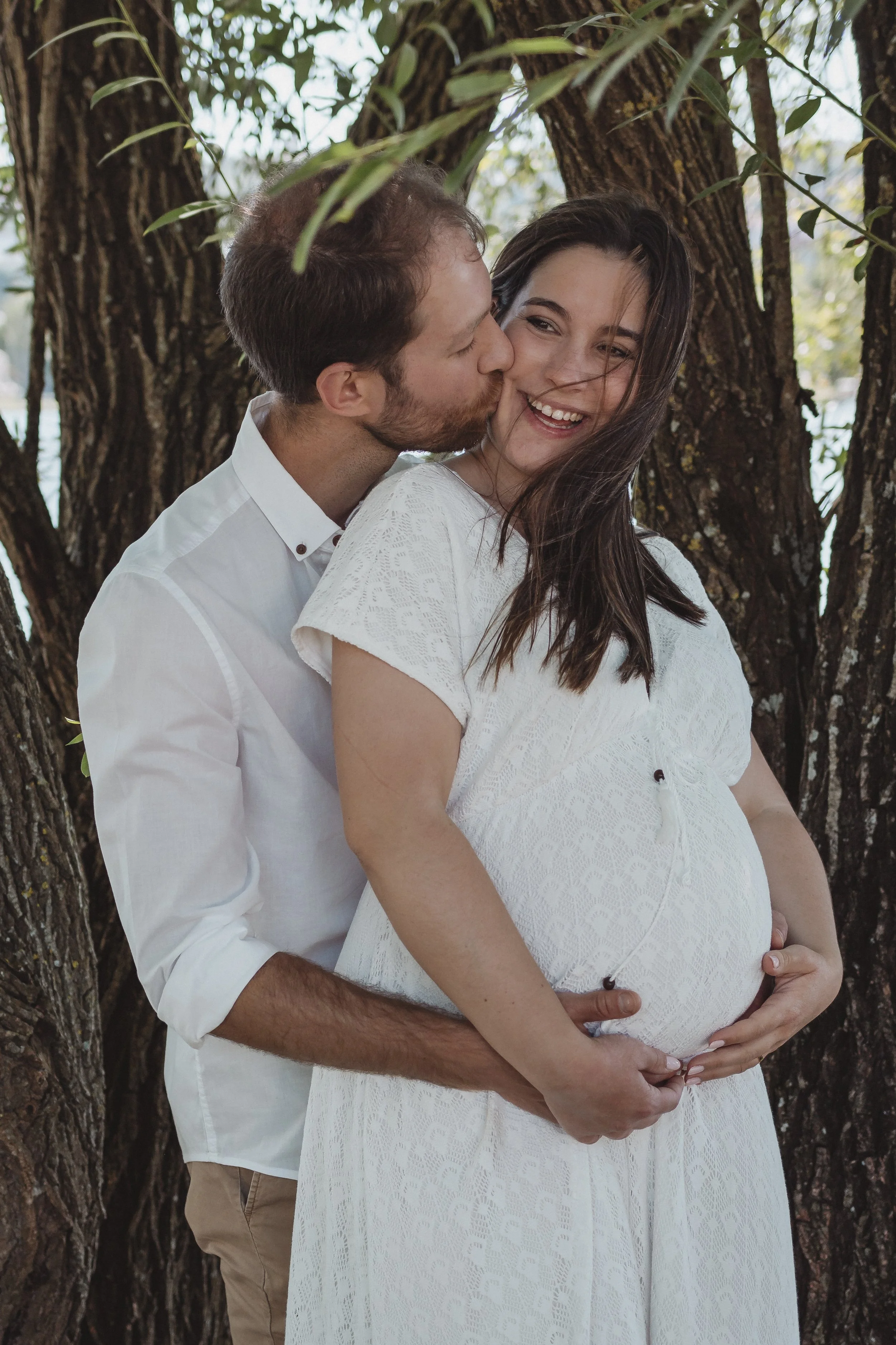 Homem beijando a mulher grávida, ambos sorrindo, ao ar livre, entre árvores.