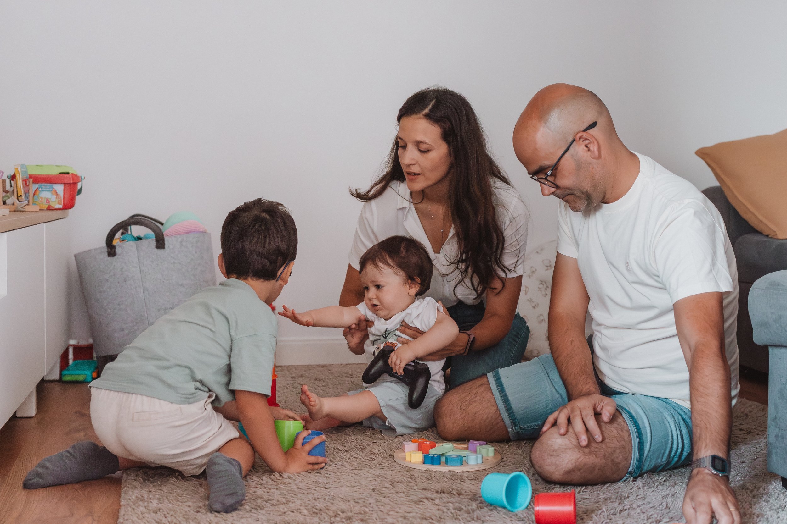 Família brincando junto no tapete da sala com brinquedos coloridos, crianças e pais interagindo.