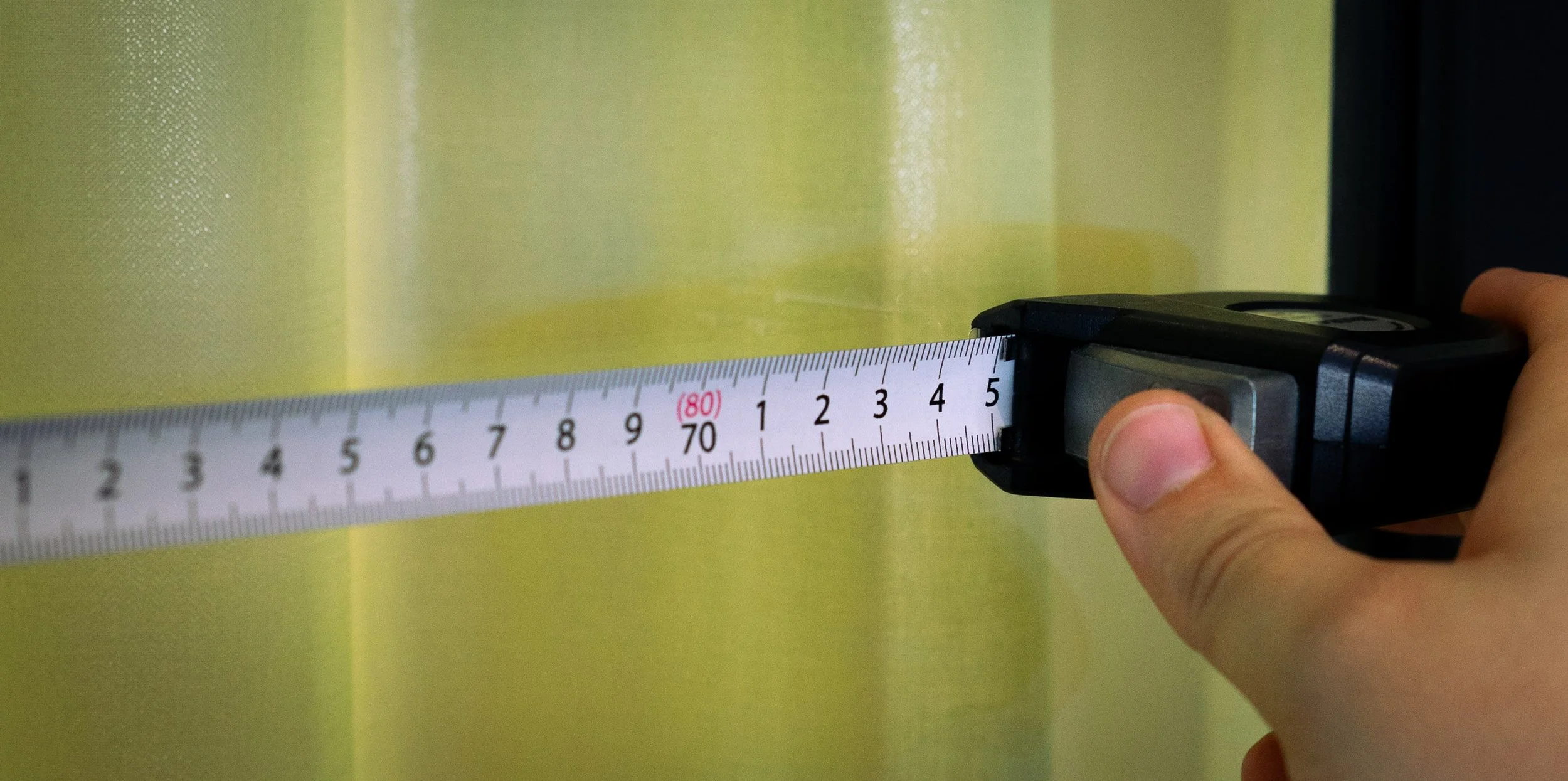 En hånd som holder en målebånd mot en gul vegg, målebåndet er utvidet til omtrent 70 centimeter.