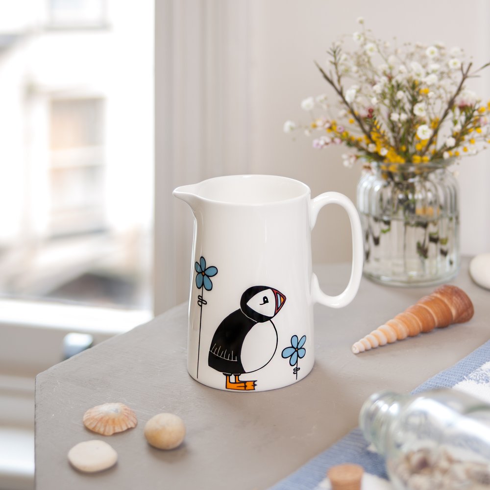  Puffin illustrated jug on table, window in background to the side, props including coastal props like shells and wildflowers 