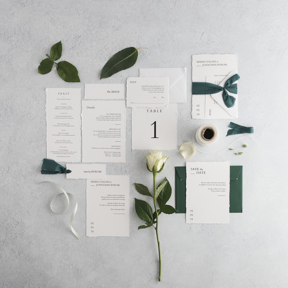  Flat-lat of complete wedding stationery suite layered with surrounding props including leaves and fresh flowers photographed upon concrete background 