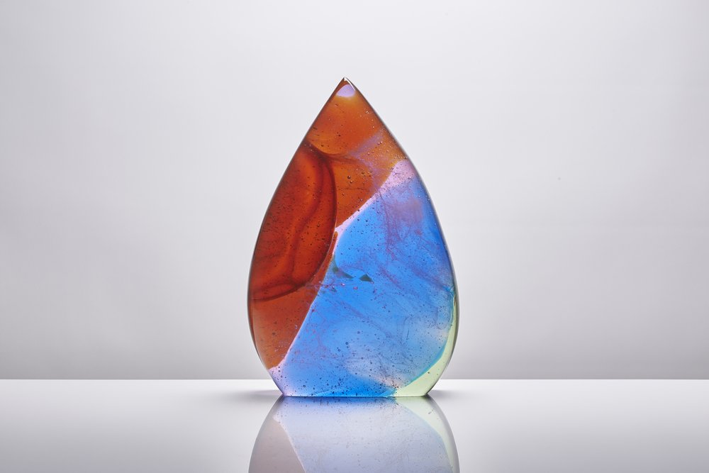  Sculptural piece of glass in vivid red and blue with intricate detailing within photographed on glossy white surface with reflection 