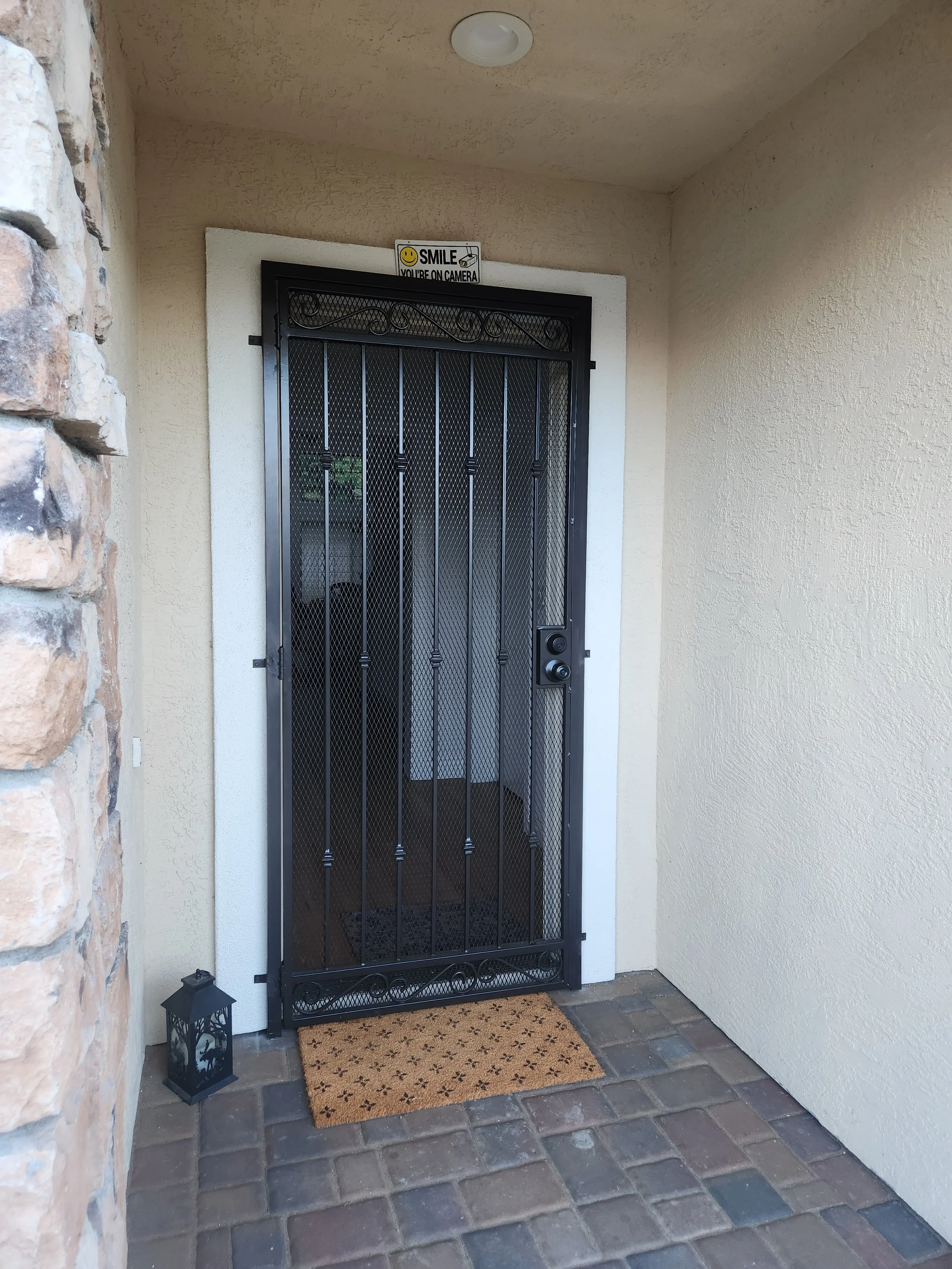 On the door of a home is the installation of a security door made of iron for better elegance. The door is black and has a brown door mat for decoration. 