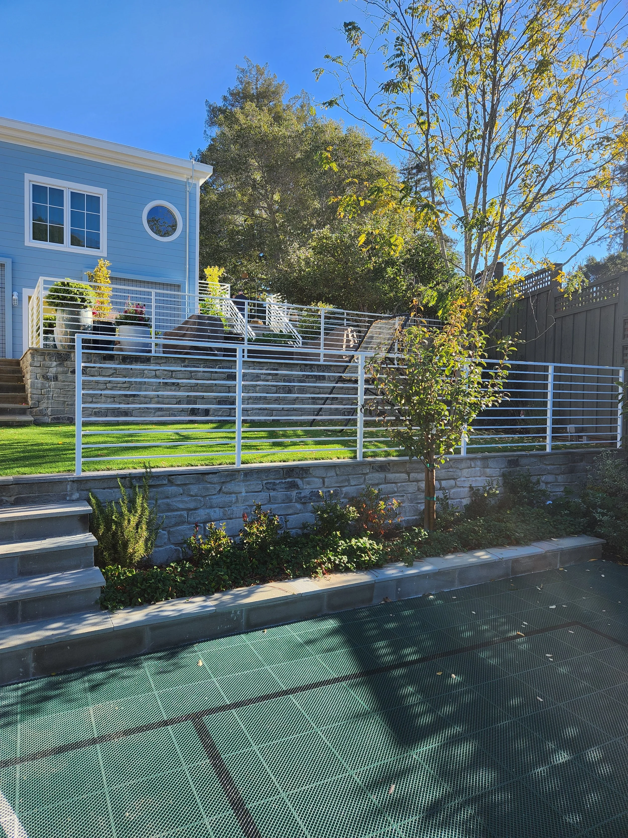 White rectangular fence for the garden of a two-story blue house separating the green area from the basketball court.