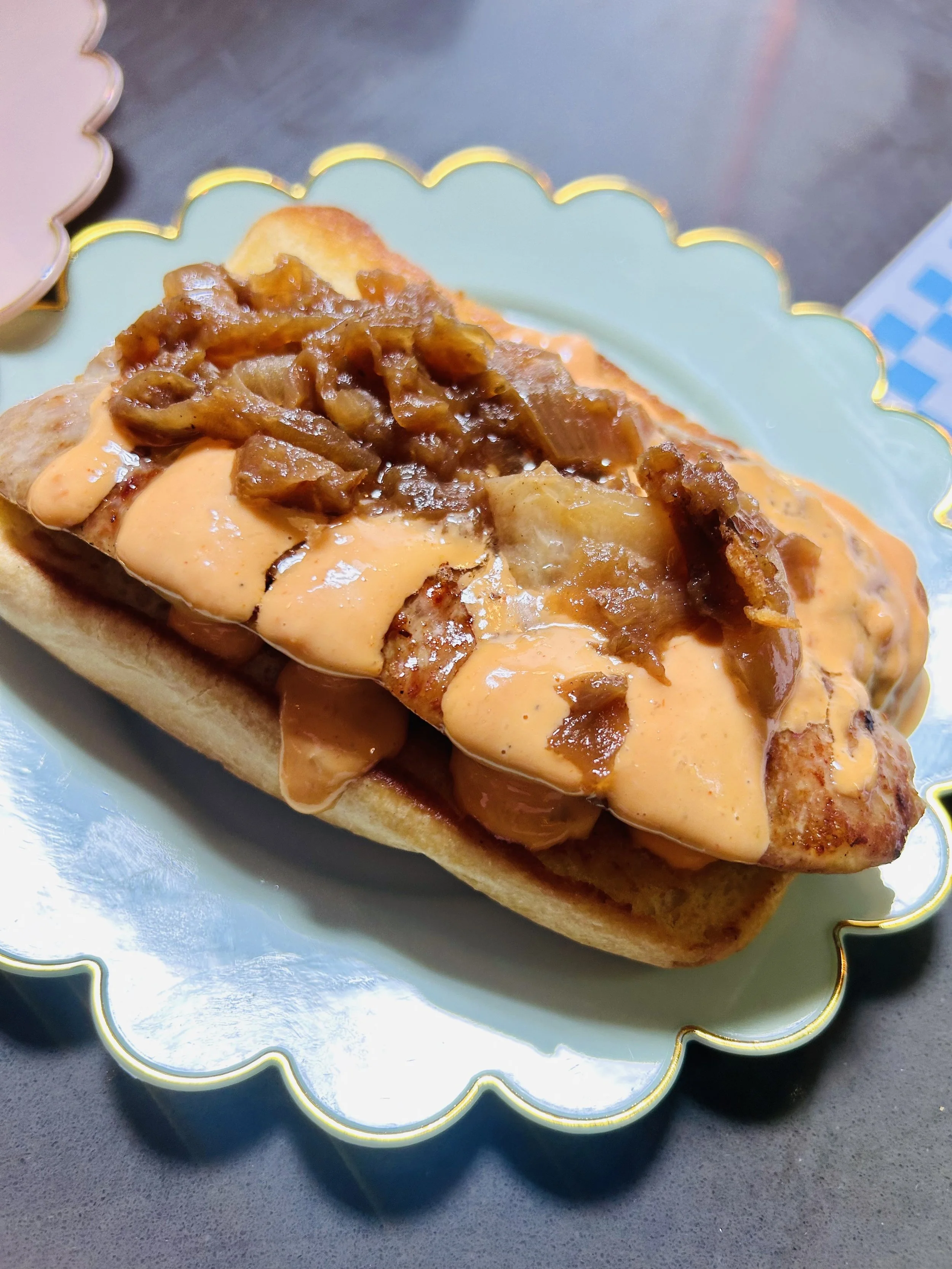 Hot dog with topped chili, onions, and drizzled sauce on a protective paper plate.