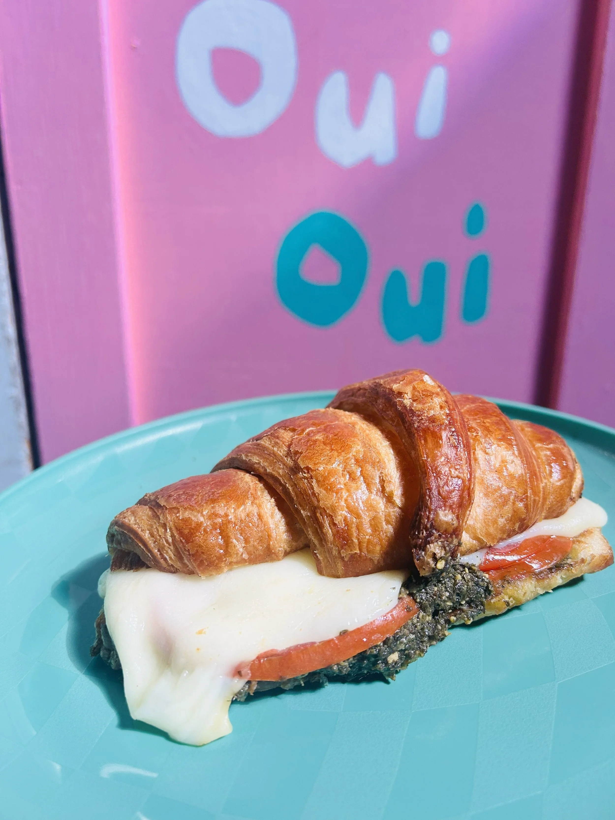 Croissant sandwich with tomato slices, melted cheese, and herbs on a turquoise plate in front of a pink sign with blue and white letters reading 'Oui Owi'.