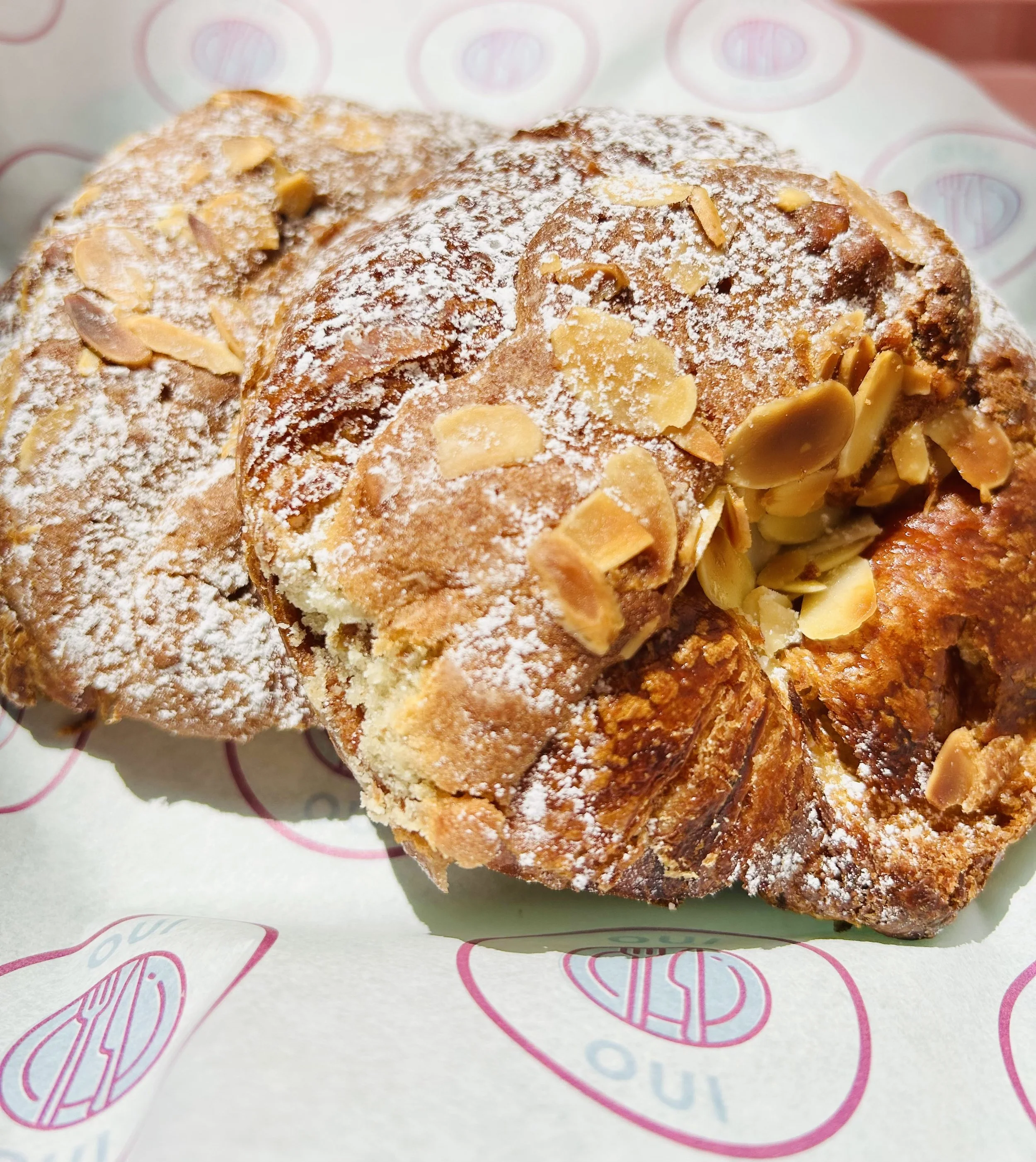 Close-up of almond croissants dusted with powdered sugar on pink and white patterned paper.