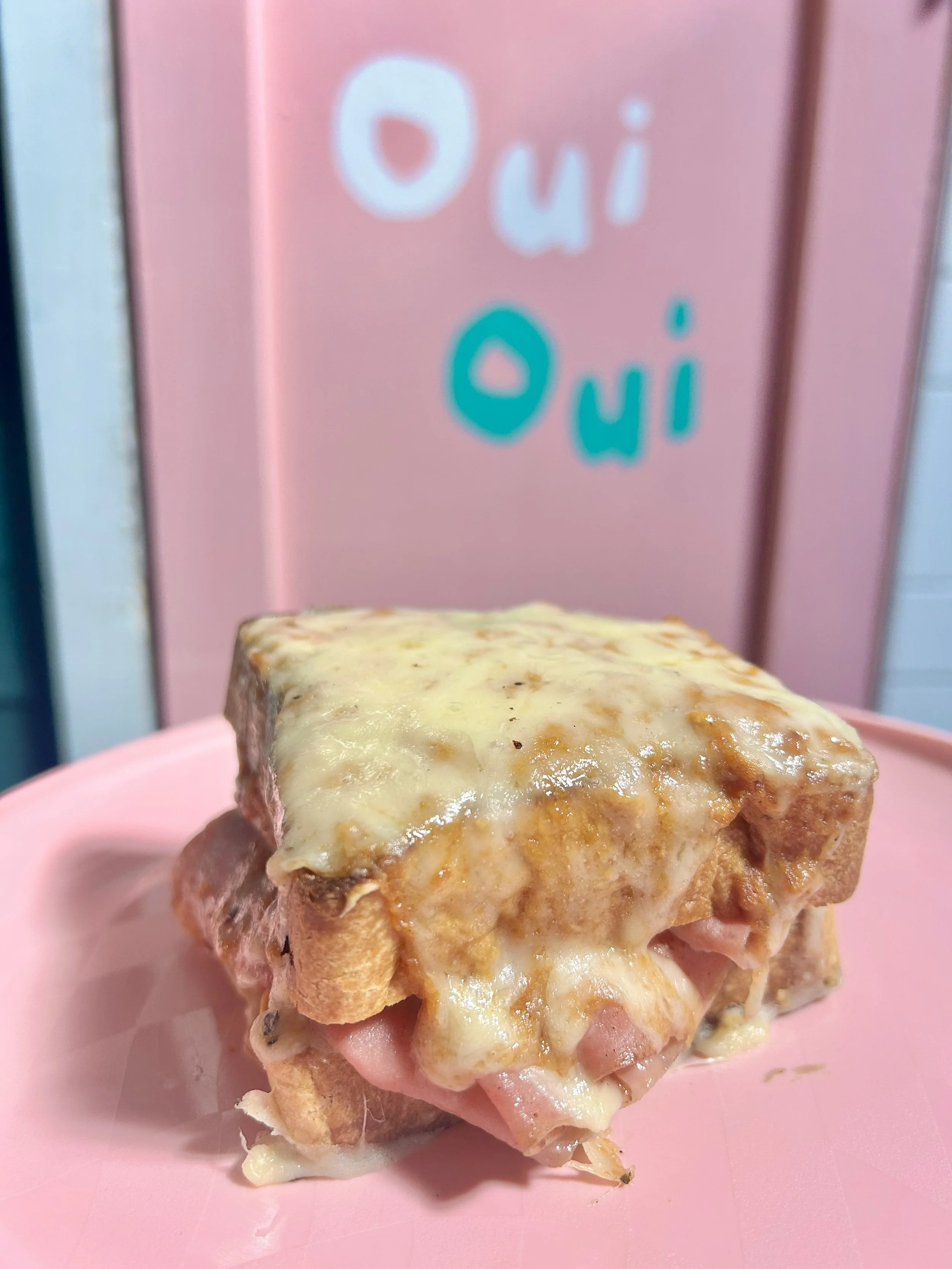 A sandwich with melted cheese on top, filled with ham, lettuce, and other ingredients, placed on a pink plate with a pink background that has the words 'Oui' written on it.